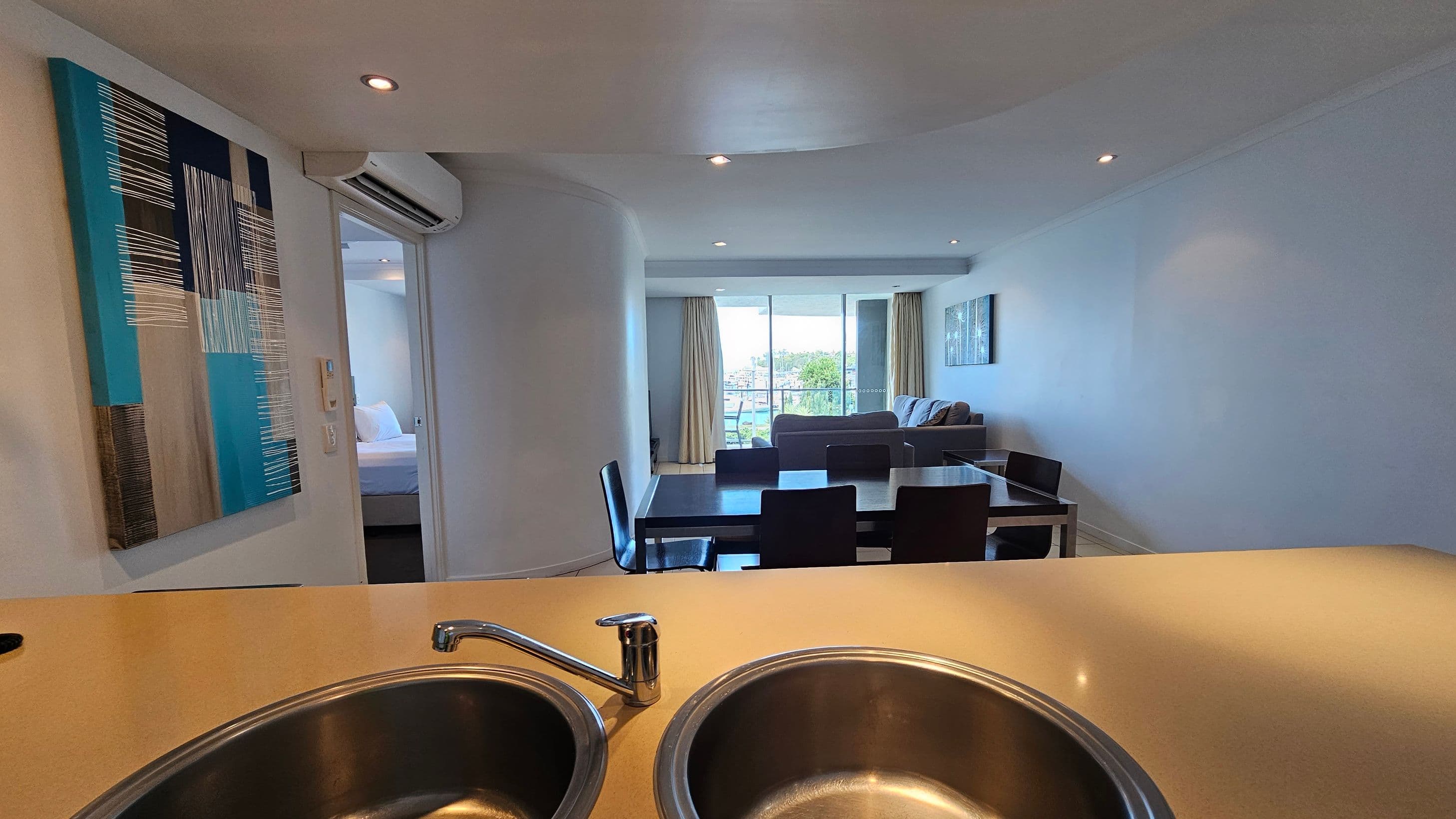 A modern interior view from a kitchen area with a double sink in the foreground. On the left, a colourful abstract painting hangs on the wall near an open doorway leading to a bedroom. In the middle, a dining table with dark chairs is visible. The background features a living room with a sofa and a large window with curtains, offering a view of trees and buildings outside. The ceiling has recessed lighting.