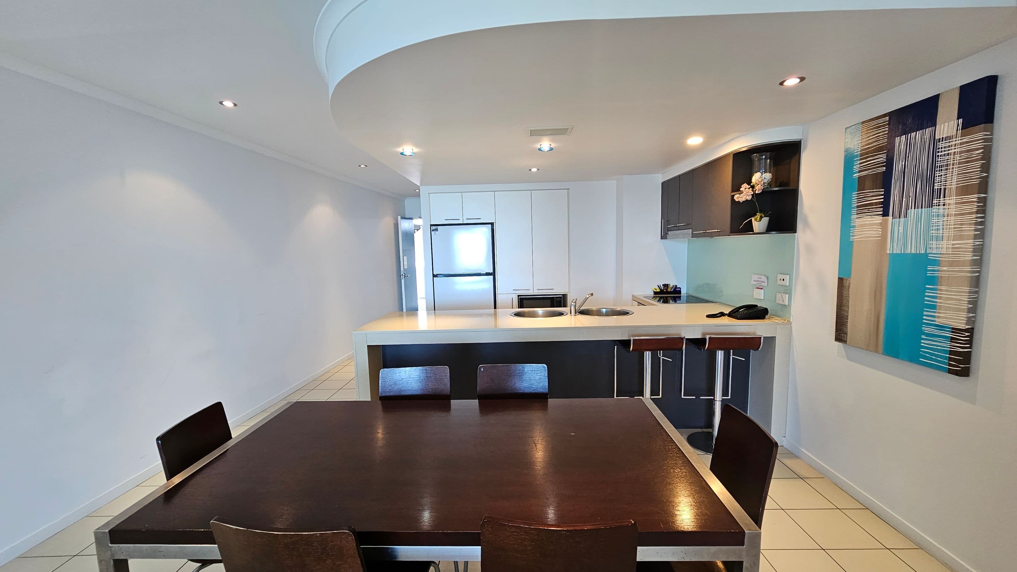 A modern kitchen and dining area with a dark wooden dining table and chairs in the foreground. The kitchen features a white countertop with two sinks, bar stools, and a phone. On the right wall, there's a colourful abstract painting. The background includes white cabinets, a stainless steel fridge, and a small plant on a shelf. The floor is tiled, and the ceiling has recessed lighting.