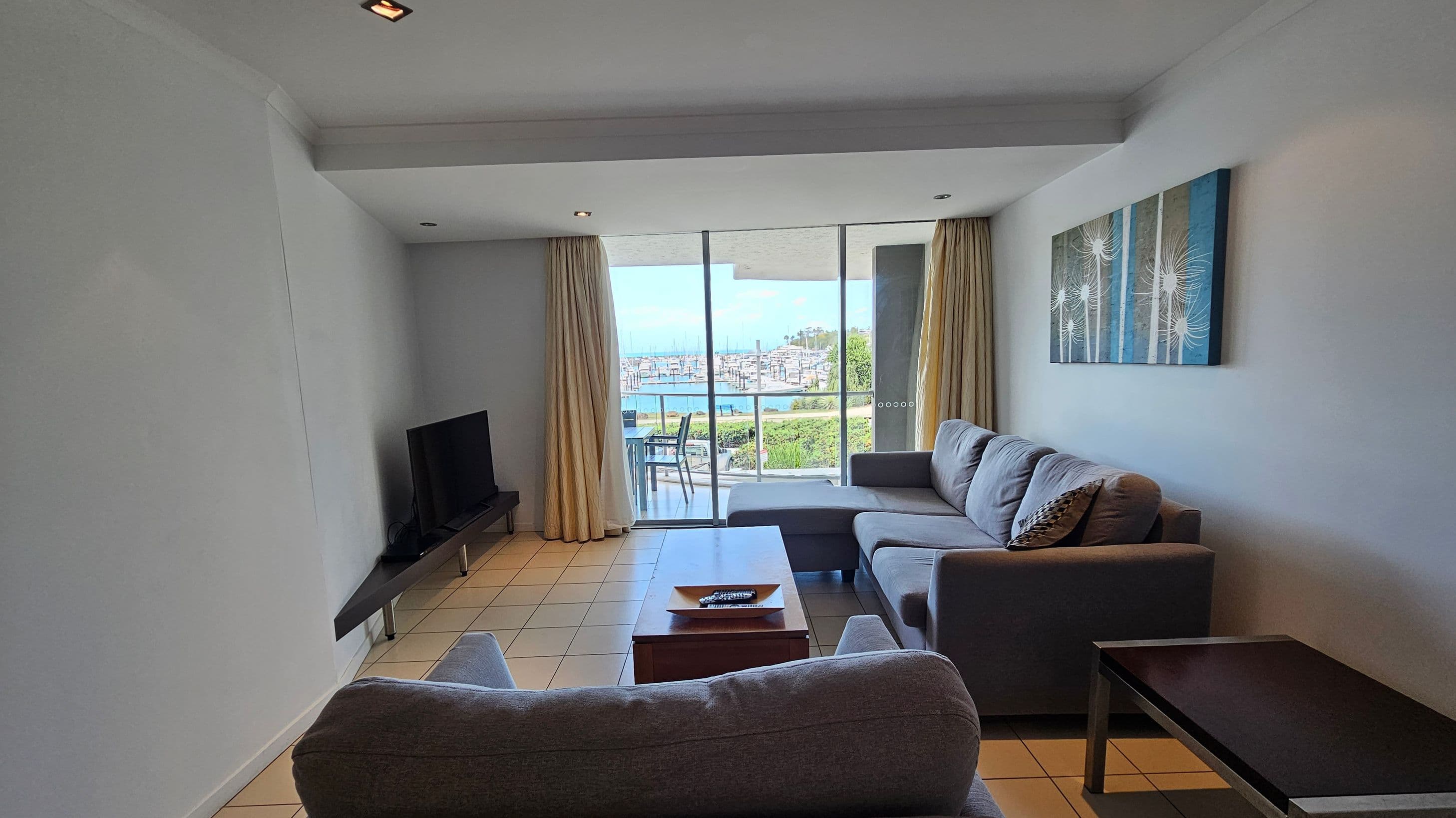 A living room with a grey sofa set on the right, a wooden coffee table in the centre, and a flat-screen TV on a stand to the left. The floor is tiled. A large glass sliding door at the back opens to a balcony with a view of a marina. Beige curtains frame the door. A piece of abstract art hangs on the right wall. The ceiling is white with recessed lighting.