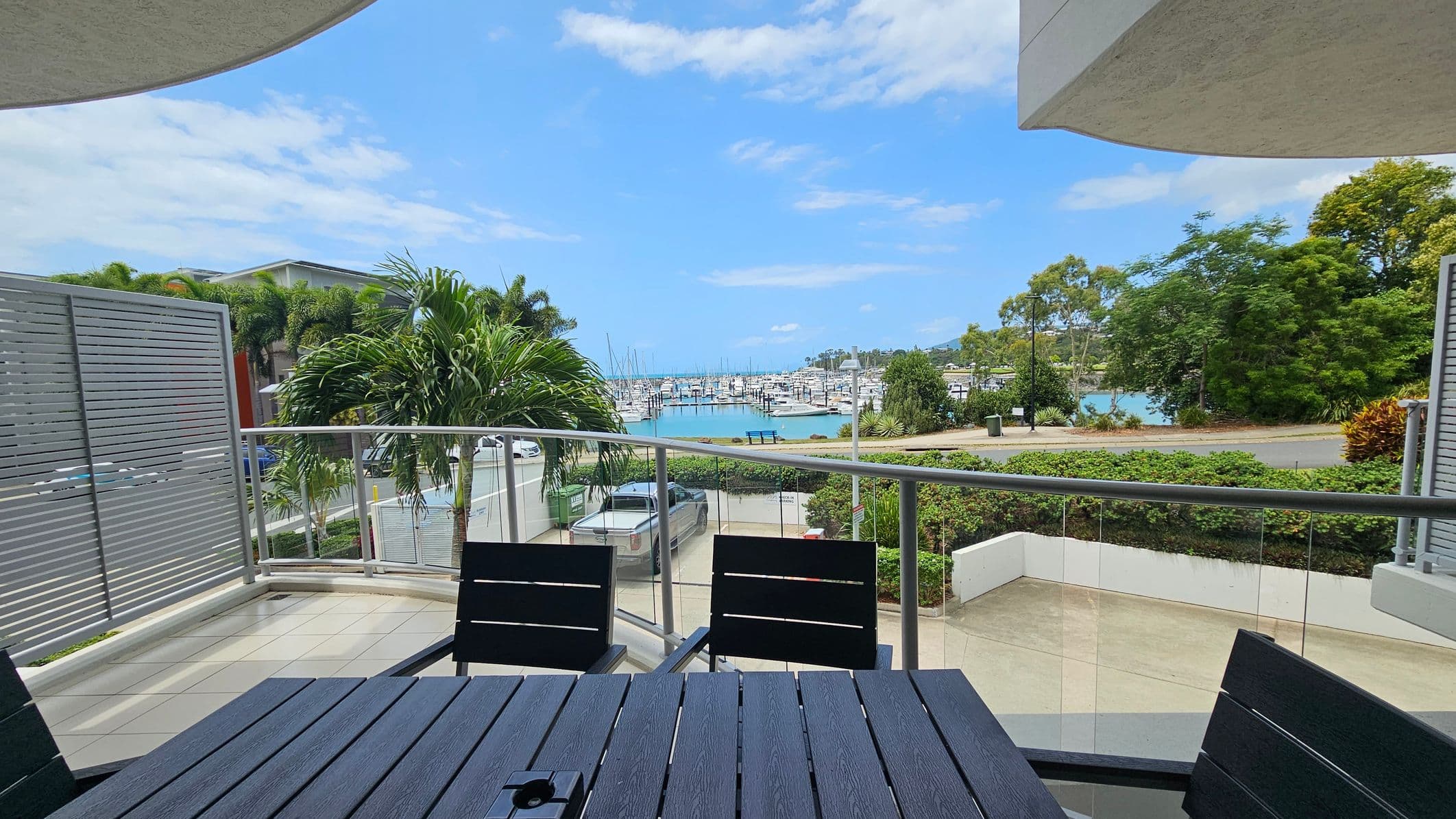 A balcony view with a dark wooden table and chairs in the foreground. The balcony has a glass railing overlooking a marina filled with numerous boats. To the left, there is a privacy screen and a palm tree. The background features a road, greenery, and more trees, with a clear view of the water and boats. The sky is blue with some clouds.