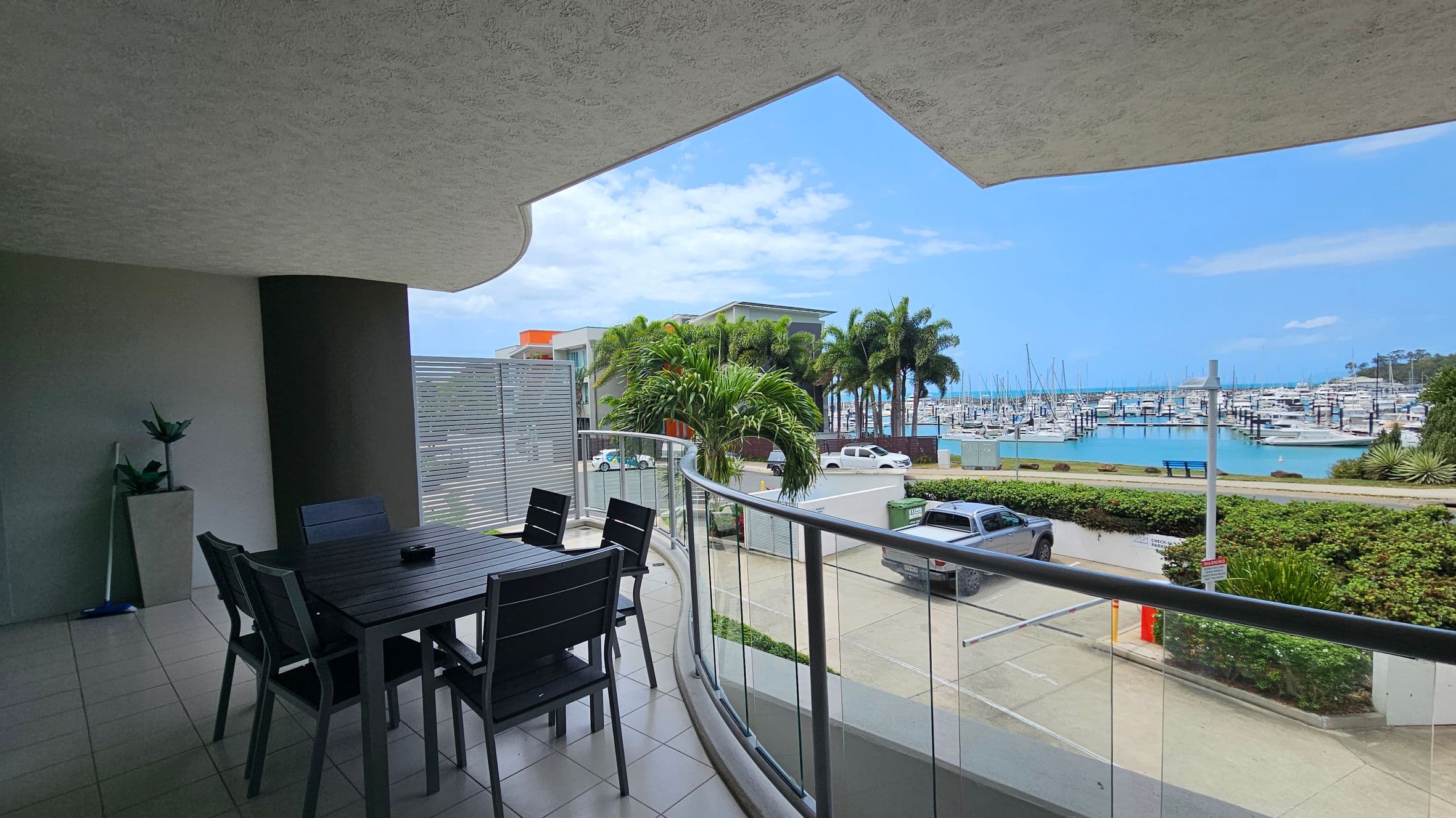 A covered balcony with a tiled floor features a dark dining table and chairs on the left. A potted plant is placed against the wall. The balcony has a glass railing overlooking a marina with numerous boats docked. In the background, there are palm trees, a few modern buildings, and parked vehicles. The ceiling is textured and curves above the seating area.
