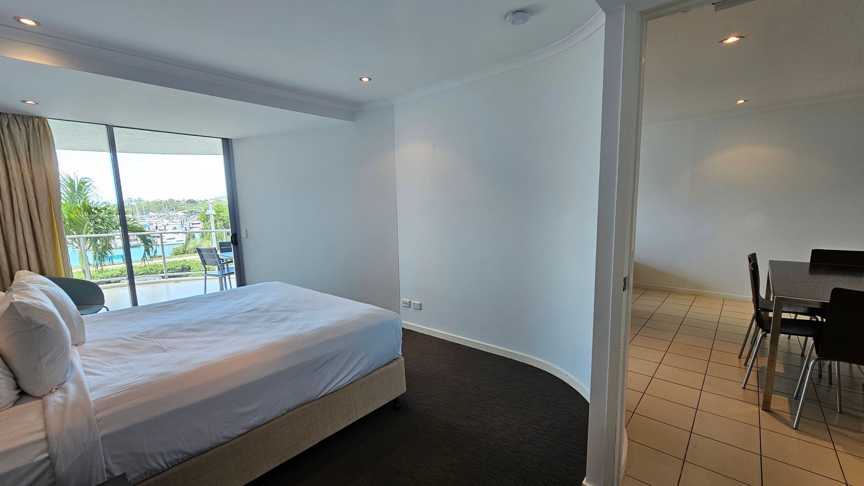 A hotel room with a bed on the left, covered in white linens, next to a large window with a view of a balcony, palm trees, and a marina. The floor is dark carpet. On the right, through an open doorway, is a dining area with a tiled floor and a table with chairs. The walls are light-coloured, and the ceiling has recessed lighting.