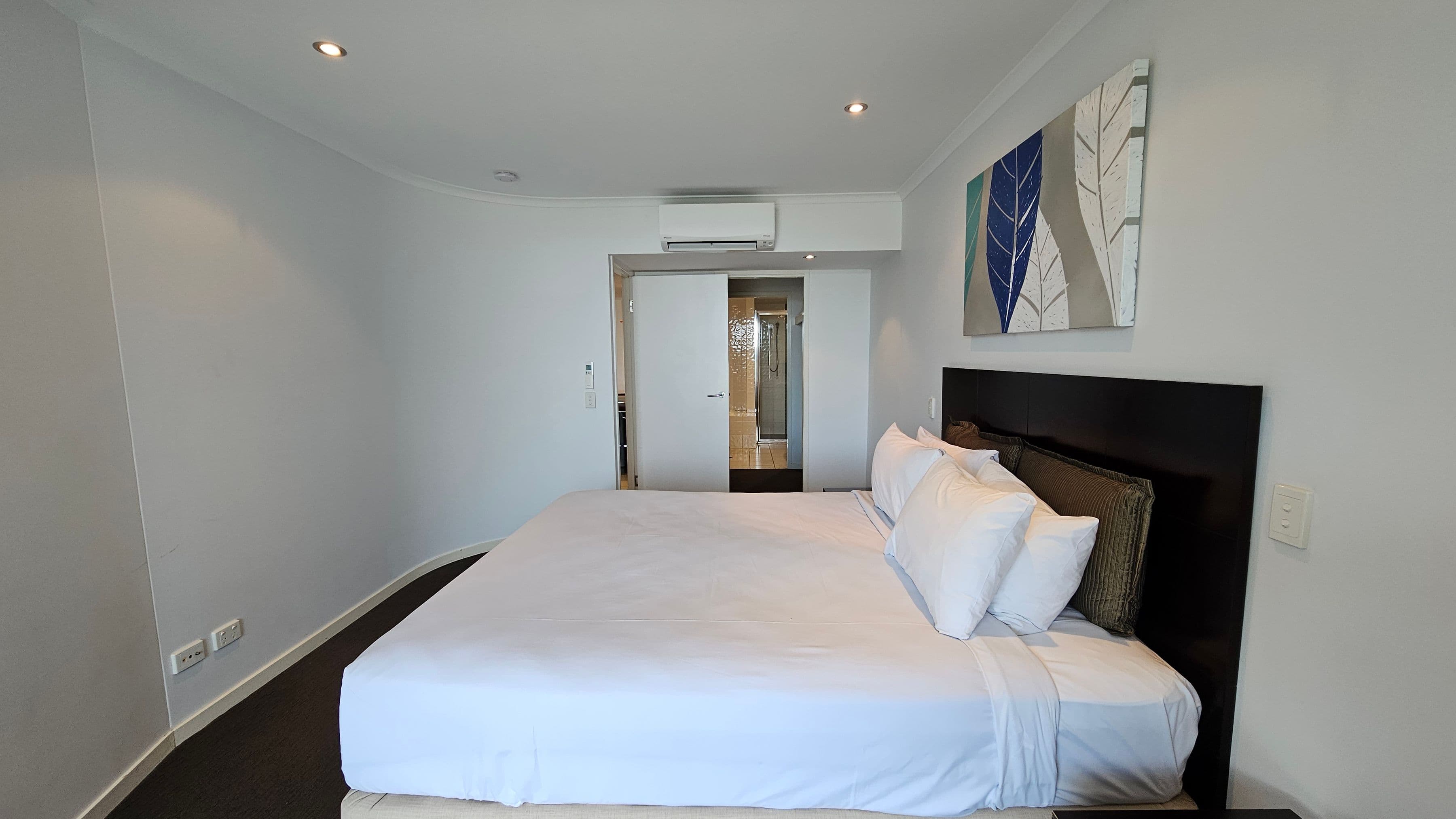 A bedroom with a large bed in the centre, covered in white linens and several pillows. Above the bed is a painting featuring leaf designs in blue and green. The walls are light grey, and the floor is carpeted in dark grey. On the left, there are electrical outlets near the floor. At the far end, a sliding door leads to a bathroom with visible shower tiles. The ceiling has recessed lighting and an air conditioning unit above the door.