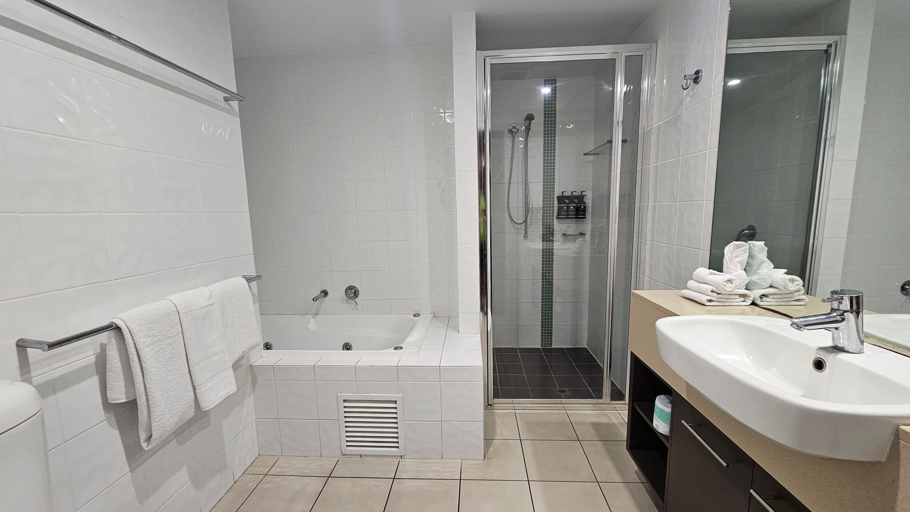 A bathroom with white tiled walls and a beige tiled floor. On the left, there's a towel rack with white towels above a bathtub. In the centre, a glass shower enclosure with a black tiled floor and a showerhead. On the right, a white sink with a chrome tap is set in a wooden vanity. The countertop holds neatly folded white towels. A mirror is mounted above the sink.