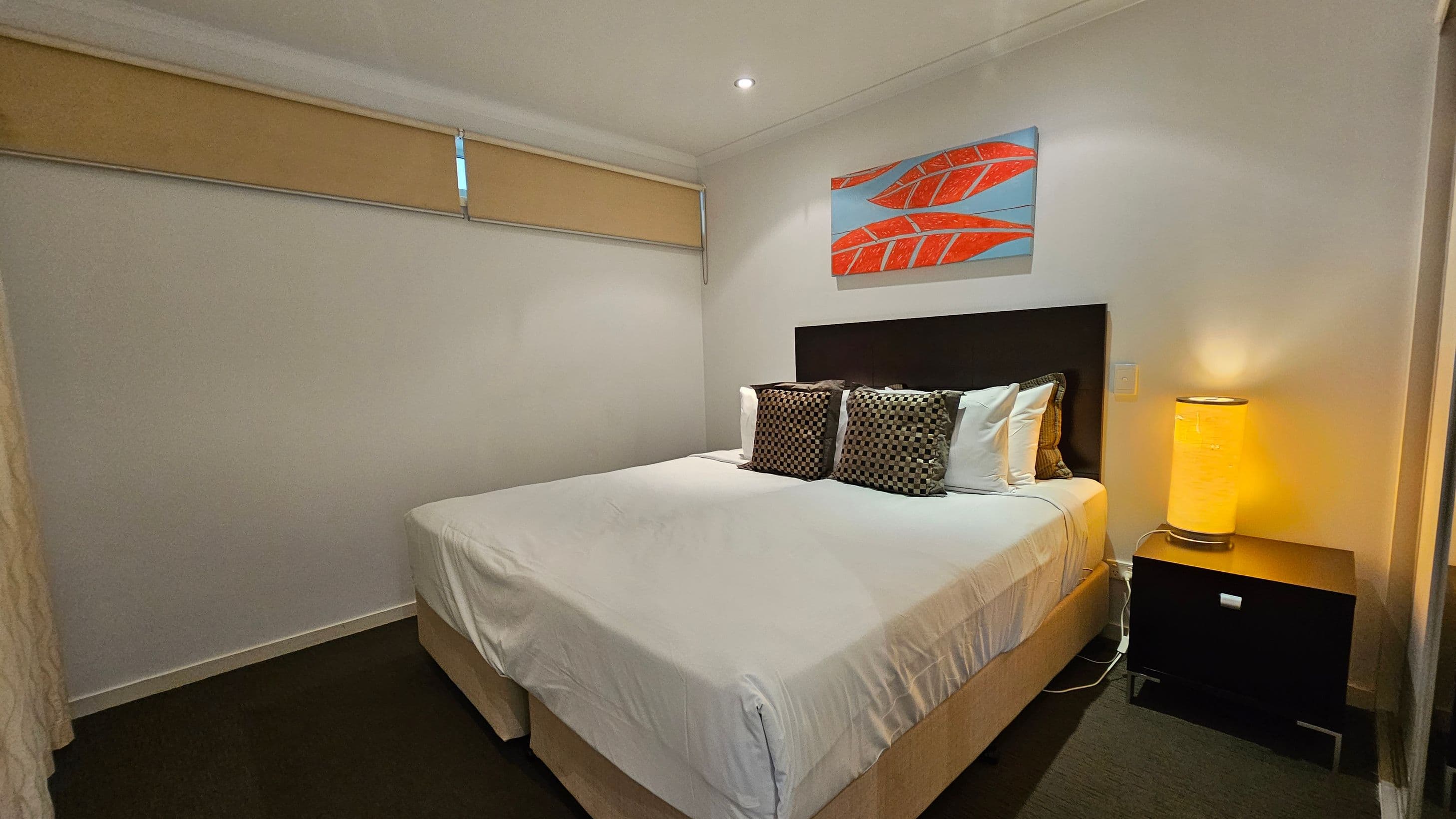 A bedroom with a bed in the centre, covered in white linens and adorned with patterned cushions. Above the bed is a painting featuring red leaves on a blue background. To the right, a bedside table holds a glowing cylindrical lamp. The left wall has a narrow window with a beige blind. The floor is carpeted in dark grey, and the ceiling is white with a recessed light.