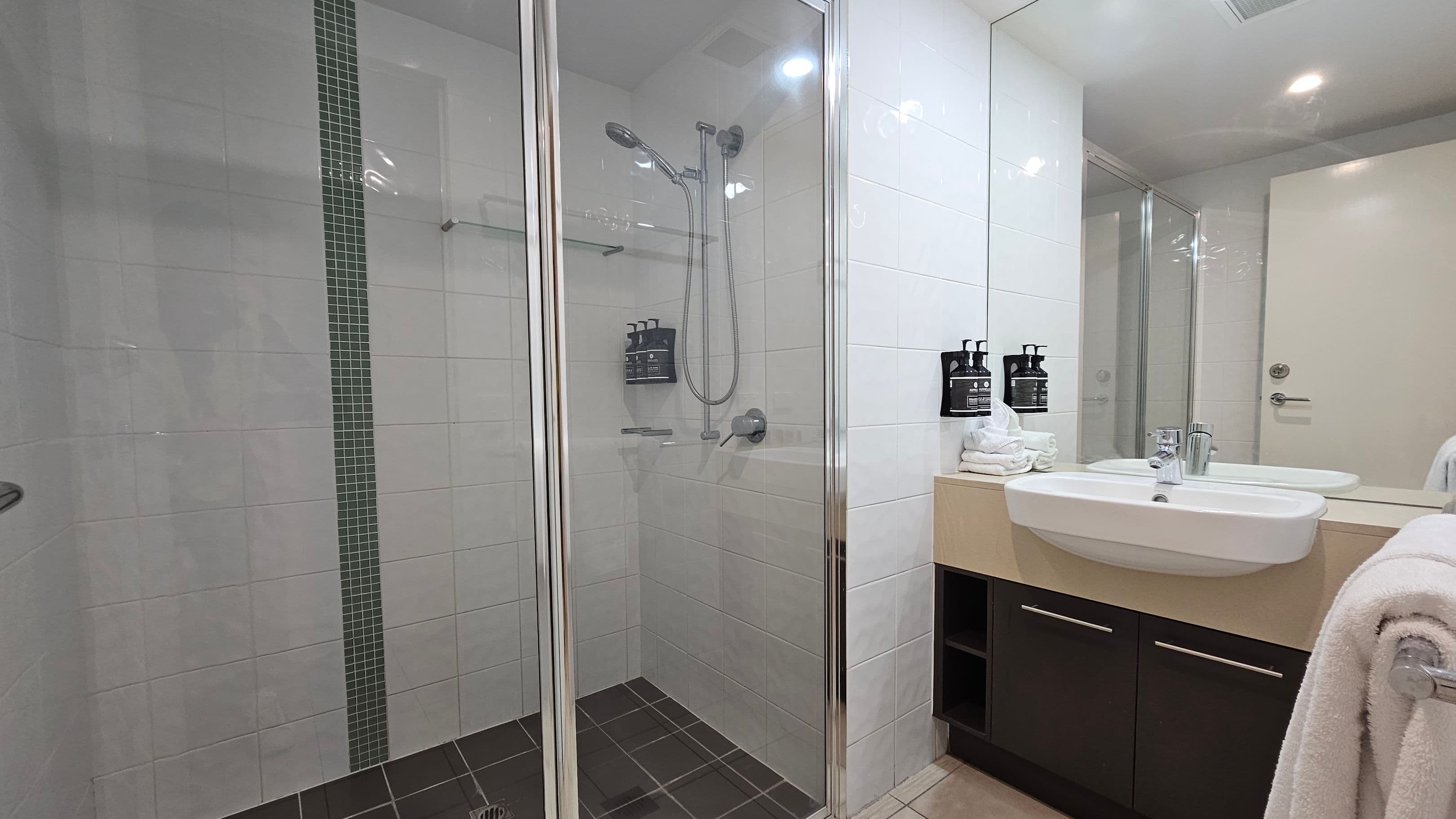 A modern bathroom with a glass-enclosed shower on the left, featuring a handheld showerhead and wall-mounted dispensers. The shower has white tiles with a vertical strip of smaller dark tiles. On the right, a white sink is set into a beige countertop with black cabinets below. Above the sink is a large mirror reflecting the room. A stack of white towels and more dispensers are on the counter. The floor is tiled, and a towel hangs on a rail to the right.