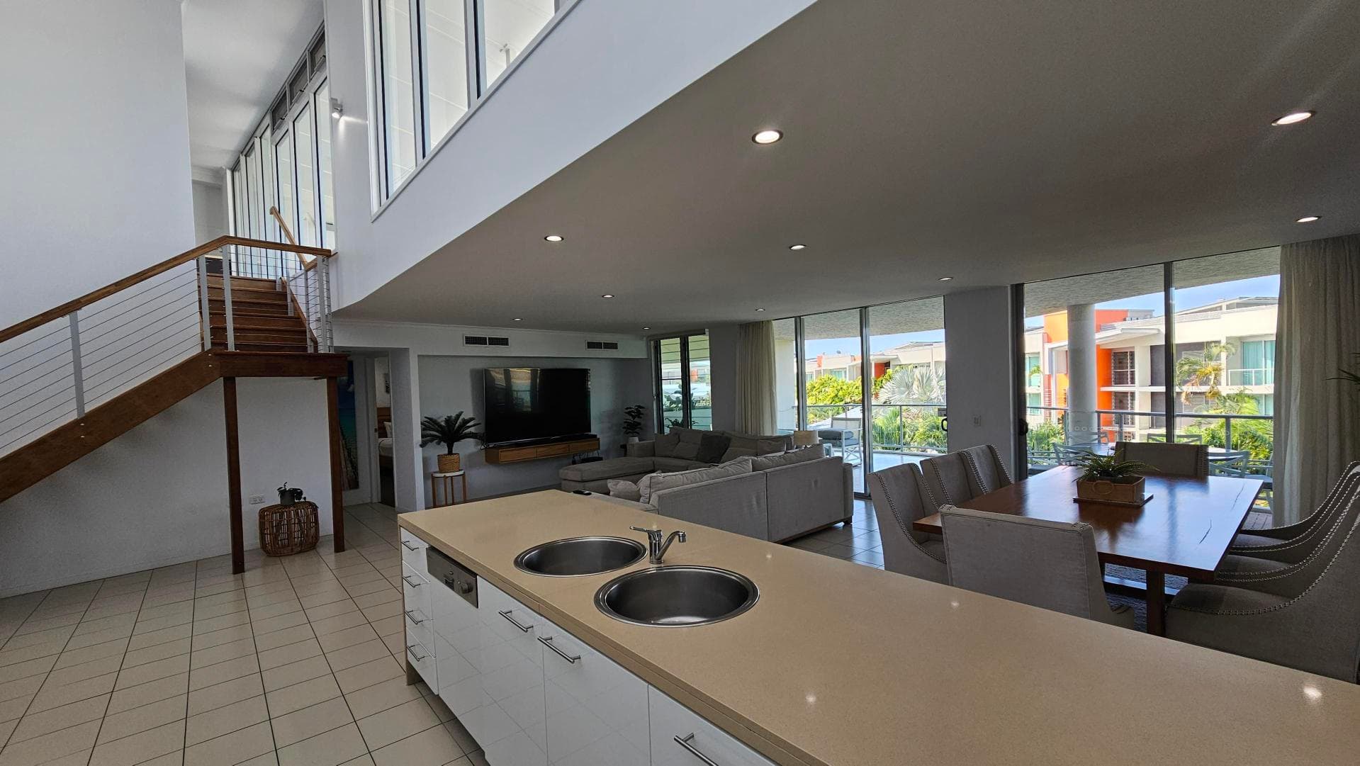 Spacious open-plan living area with a tiled floor. On the left, there's a large grey sectional sofa on a dark rug, facing a wall of windows with sheer curtains, offering a view of greenery outside. In the centre, a dining table with white chairs is positioned near a wall with a beach-themed painting. On the right, a modern kitchen features white cabinets, a beige countertop, and a sink. The ceiling is high with recessed lighting.