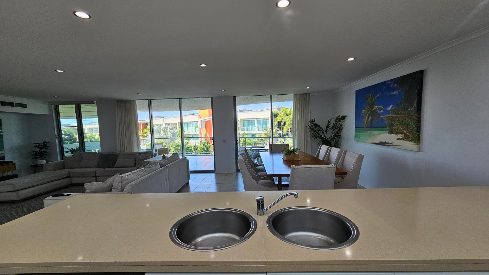 A modern open-plan living space with a kitchen island in the foreground featuring a double sink. To the left, there's a large grey sectional sofa facing a glass sliding door that opens to a balcony with greenery outside. On the right, a wooden dining table with white chairs is set against a wall adorned with a beach-themed painting. The ceiling is fitted with recessed lighting, and the floor is tiled.