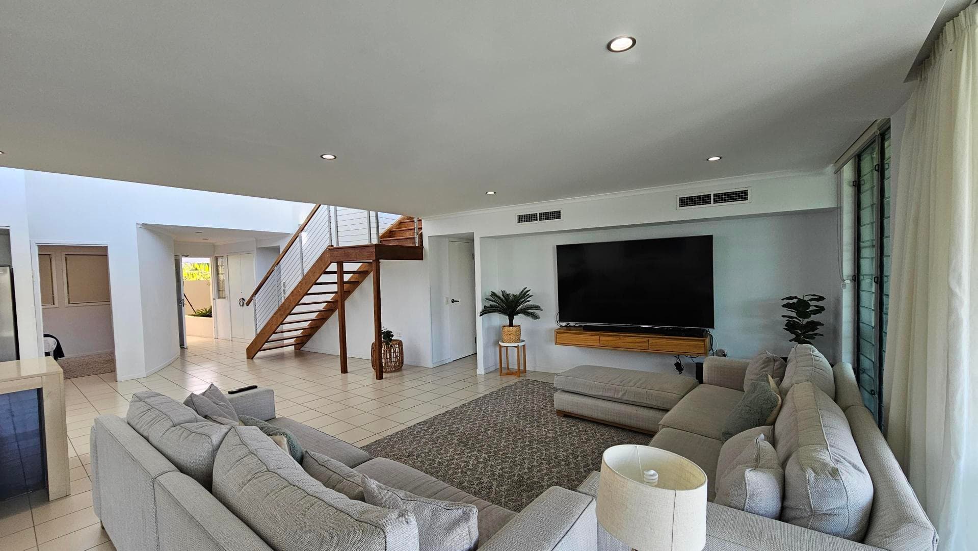 A modern living room with light-coloured tiled flooring. On the right, a large flat-screen TV is mounted on the wall above a wooden console. Below, a grey sectional sofa faces the TV, with a small round table beside it. A patterned rug lies on the floor. To the left, a wooden staircase with metal railings leads upstairs. Large windows with sheer curtains are on the far right, allowing natural light in. A potted plant is placed near the TV. The ceiling is white with recessed lighting.