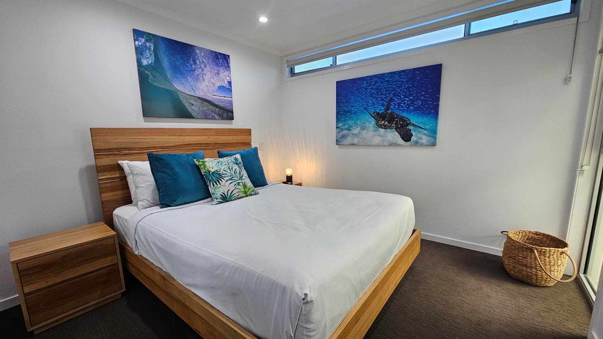 A bedroom with a wooden bed frame and white bedding, accented by blue and green pillows. On the left, there's a wooden bedside table. Above the bed, a large artwork of a wave is displayed. On the right wall, another artwork features a sea turtle underwater. A woven basket sits on the floor near a glass door. The room has a dark carpet and a narrow window near the ceiling.