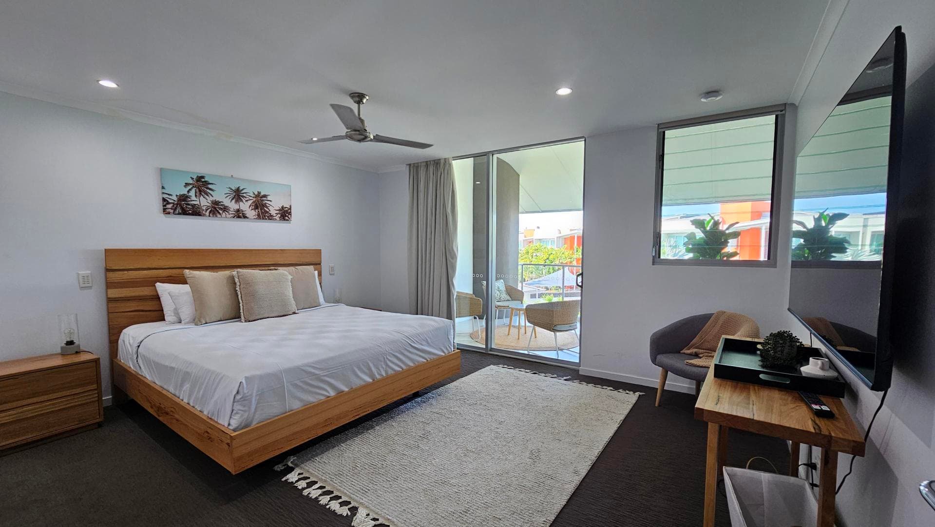 A small, minimalist bedroom with white walls and a light grey patterned carpet. On the left, a wooden cabinet is partially visible. A single bed with a white duvet and a dark blue throw is against the left wall, featuring a decorative pillow with a leaf pattern. Above the bed, a beach-themed canvas with starfish hangs. Two windows with closed blinds are on the back wall. A tall, freestanding mirror leans against the right wall. The ceiling has recessed lights.