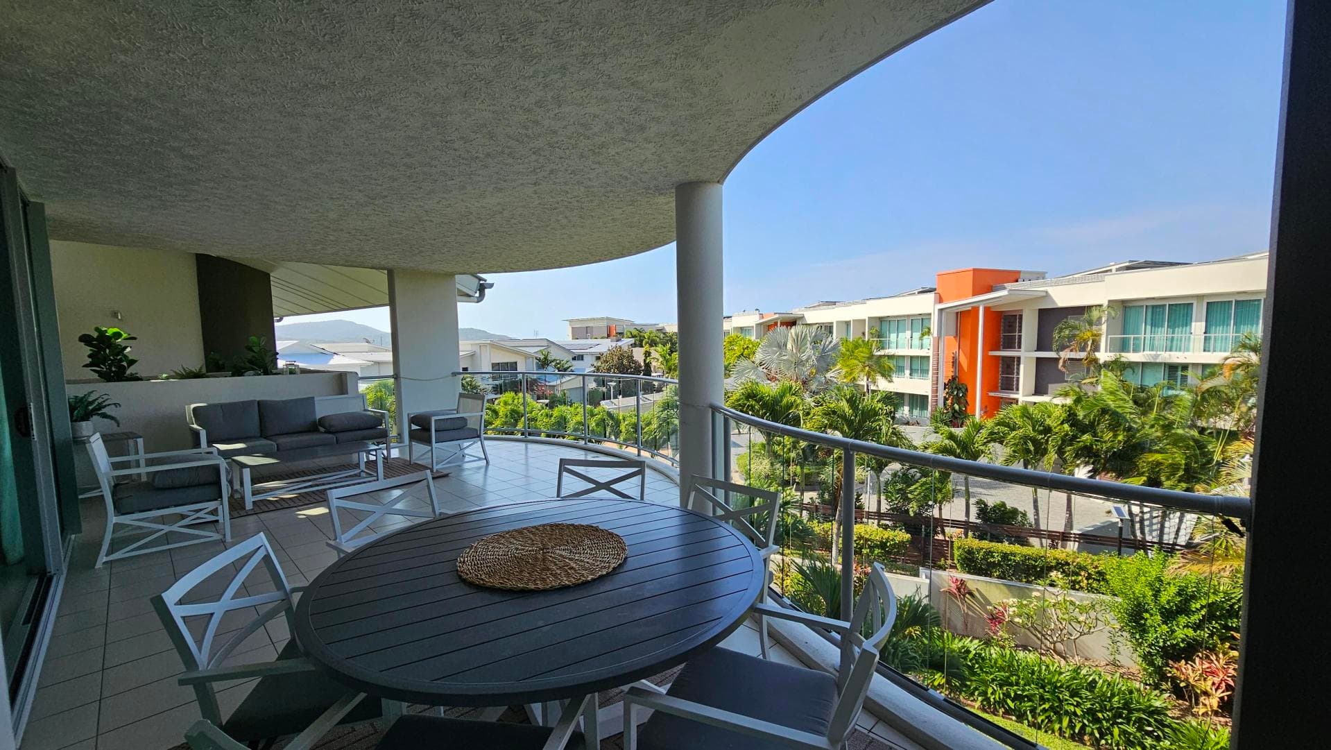 A spacious balcony with a round table and chairs in the foreground. To the left, there is a seating area with a sofa and armchairs, along with potted plants. The balcony has a glass railing, offering a view of a modern building with orange accents and lush greenery in the background. The ceiling is textured, and the floor is tiled.