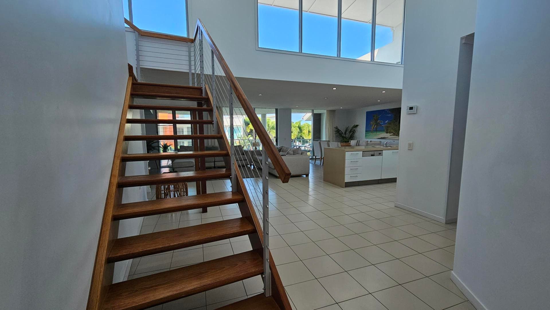 A modern interior with a wooden staircase on the left, featuring metal railings. The floor is tiled in white. To the right, there's a kitchen area with white cabinets and a visible dishwasher. In the background, a living area with sofas and large windows allowing natural light. The ceiling is high with recessed lighting, and a large painting is visible on the wall.