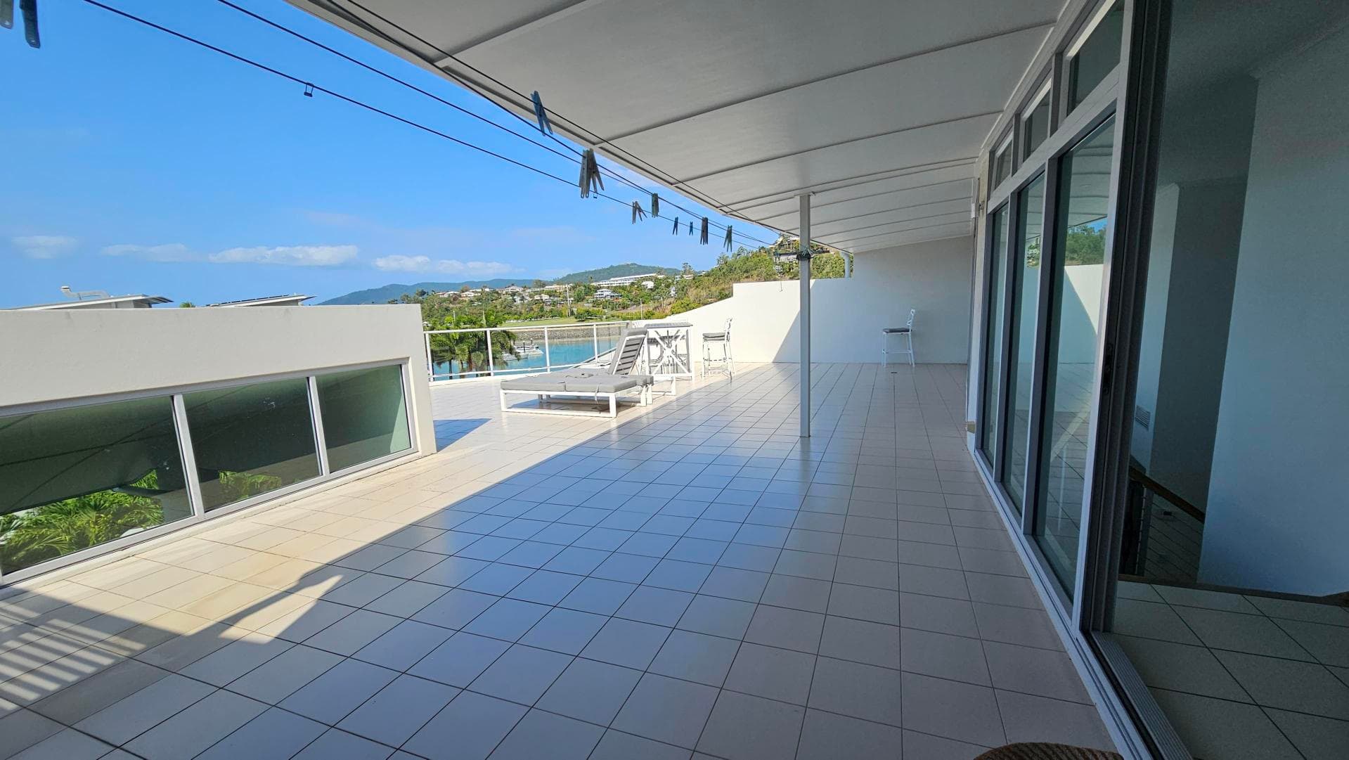 A spacious terrace with a tiled floor and a white ceiling. On the right, there are large glass doors reflecting the surroundings. In the middle, a sun lounger and a small table with chairs are positioned near a glass railing. On the left, a modern building with large windows is visible. The background features a view of greenery and distant hills. Clothes pegs hang from lines above.