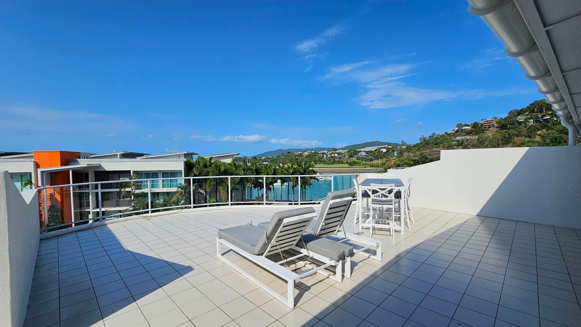 A spacious terrace with a tiled floor features two white sun loungers with grey cushions in the centre. To the right, there's a white high table with matching chairs. The terrace is bordered by a glass railing, offering views of modern buildings with orange and grey accents, palm trees, and a distant hillside. The sky is clear and blue.