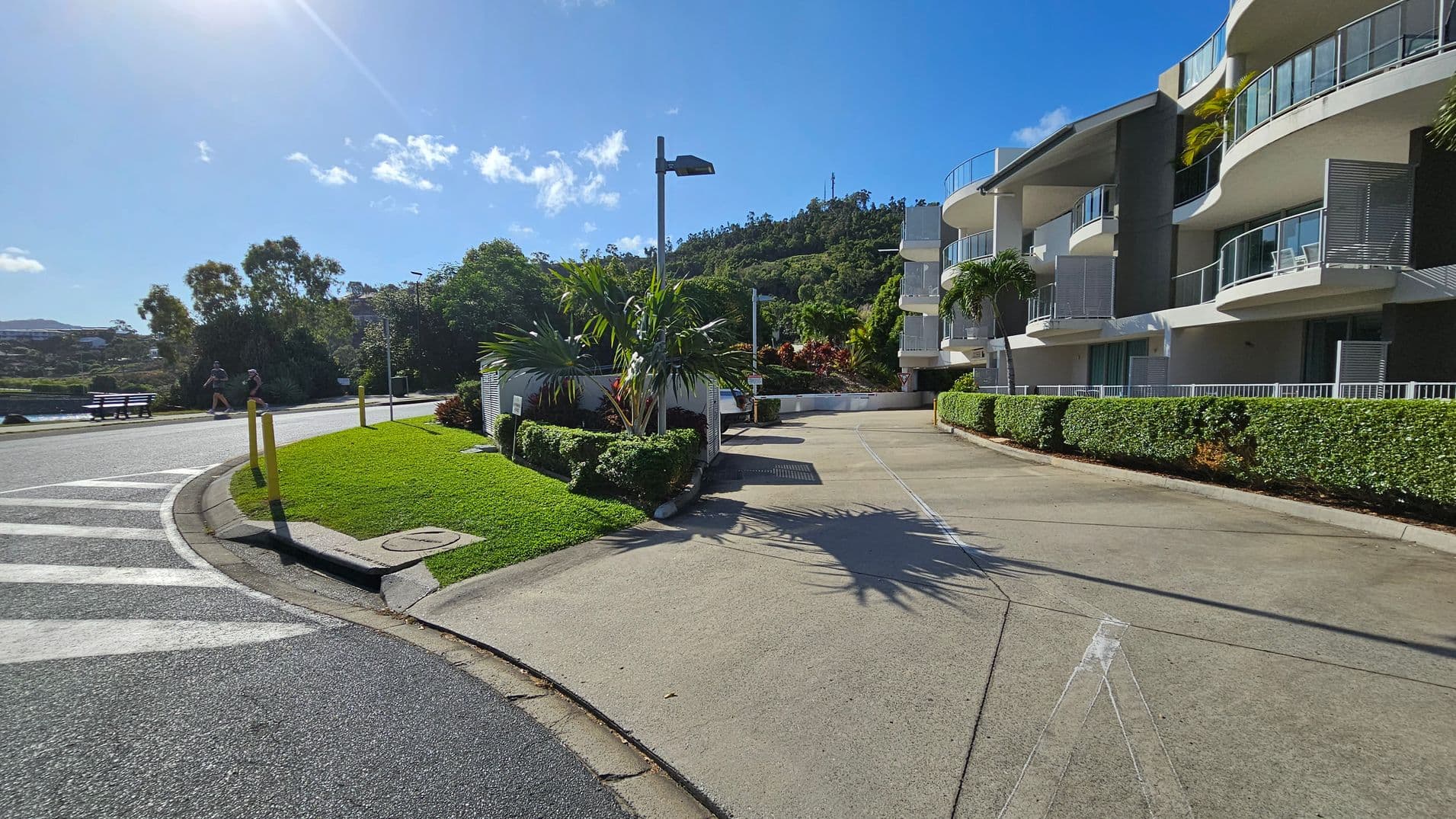 A curved road on the left leads towards a lush, green hillside in the background. On the right, a modern, multi-storey building with balconies is surrounded by well-maintained hedges and palm trees. The foreground features a small grassy area with yellow bollards and a utility box. The road surface is smooth and dark, with white markings near the kerb.