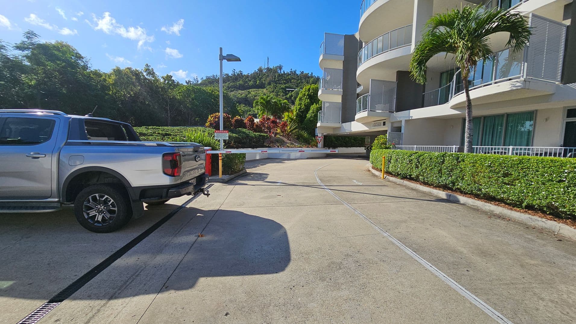 A silver pickup truck is parked on the left side of a concrete driveway. On the right, there's a modern building with curved balconies and glass windows, bordered by a neatly trimmed hedge. A palm tree stands near the building. In the background, lush green trees and bushes are visible, with a hill rising behind them. A barrier gate and a streetlamp are positioned near the centre of the image.