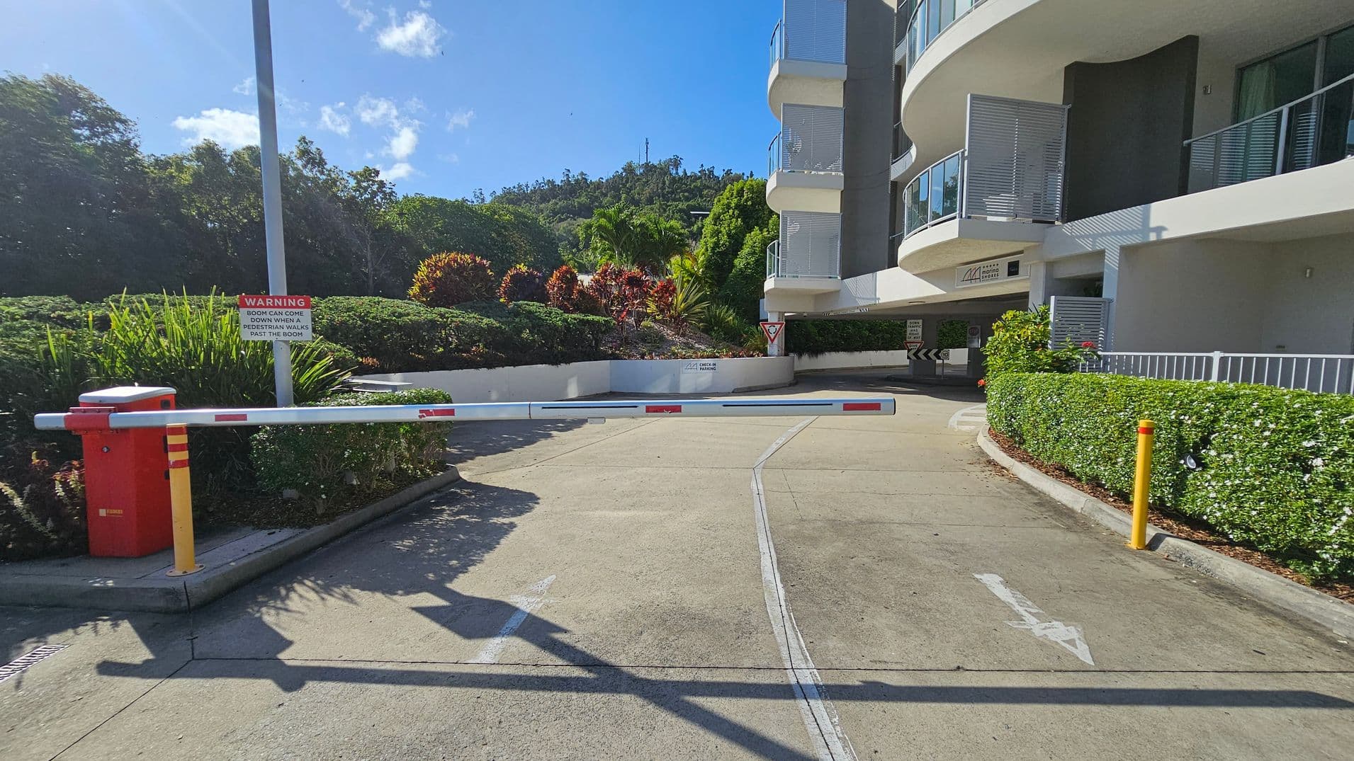 A driveway with a raised boom gate in the foreground. On the left, there's a red and yellow control box and a warning sign about the boom. Lush greenery and bushes line the left side. On the right, a modern building with curved balconies and glass railings. A hedge borders the pavement. In the background, a hill with dense trees and a clear blue sky. The ground is paved with light-coloured concrete, marked with arrows.