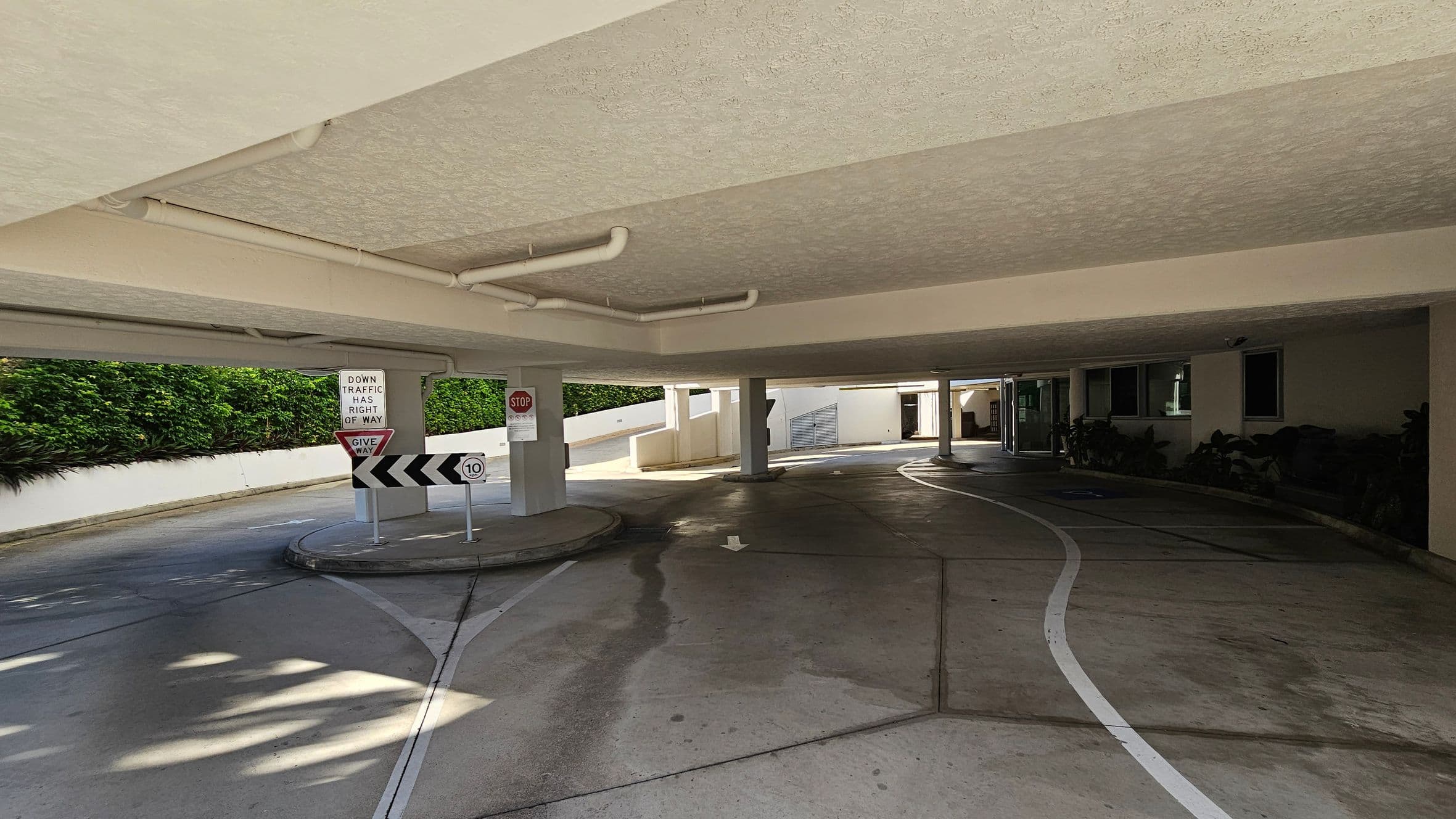 A covered parking area with a concrete floor and ceiling. On the left, there are traffic signs including "Give Way" and "Down Traffic Has Right of Way," along with a speed limit sign of 10. A stop sign is mounted on a pillar. The right side features a building with windows and some plants. The background shows a curved driveway leading out, bordered by a white wall and greenery. Pipes are visible on the ceiling.