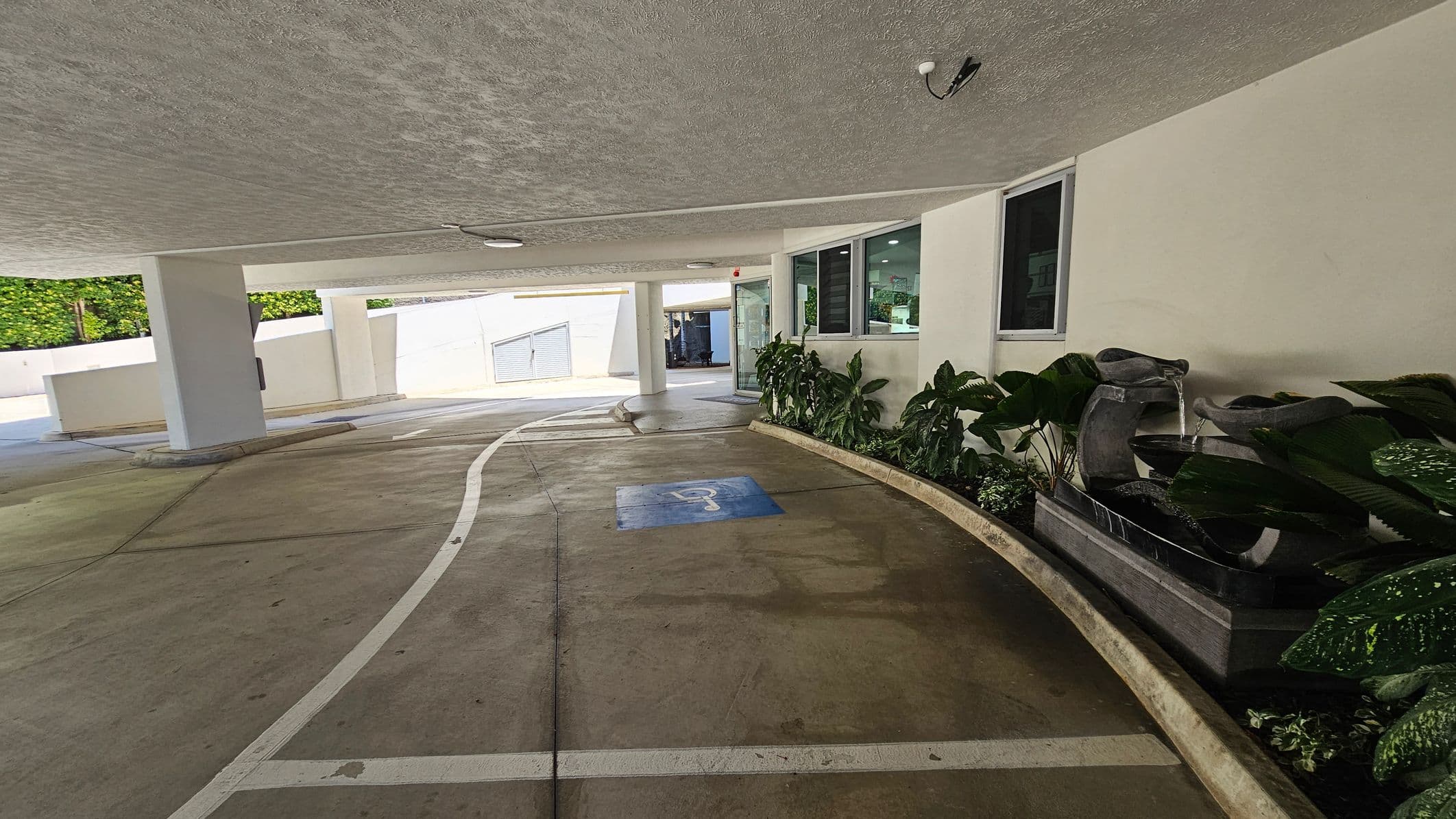 Covered parking area with a smooth concrete floor and a textured ceiling. On the right, there's a row of large green plants and a decorative water feature with cascading water. Windows are visible above the plants. A blue disabled parking symbol is painted on the floor. In the background, there are white walls and a partially open gate. Ceiling lights and a security camera are mounted above.