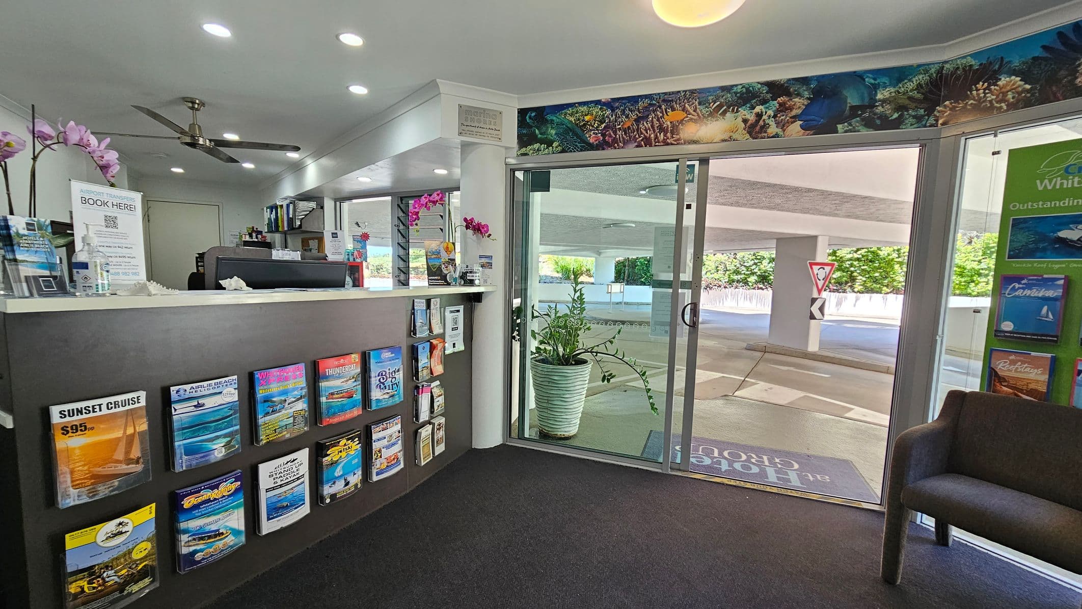 A reception area with a counter on the left displaying brochures and a sign for airport transfers. Above the counter, there are pink orchids and a ceiling fan. The floor is carpeted. On the right, a glass door leads outside, with a large potted plant nearby. Brochures are also displayed on the wall next to the door. The ceiling has recessed lighting, and a colourful marine-themed border decorates the top of the walls.