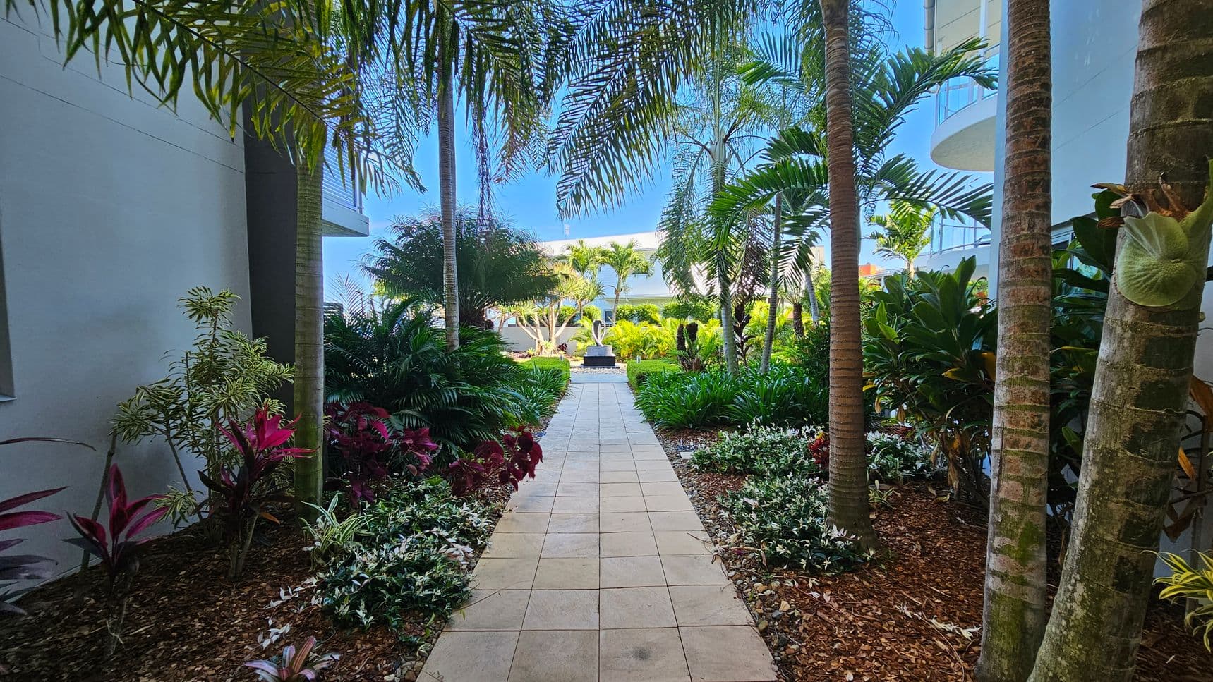 A paved walkway leads through a lush garden with various tropical plants and trees on both sides. On the left, there are red and green leafy plants, while on the right, there are dense green shrubs and palm trees. In the background, a modern building with glass balconies is partially visible, and a sculpture is centred at the end of the path. The ground is covered with mulch and small stones.