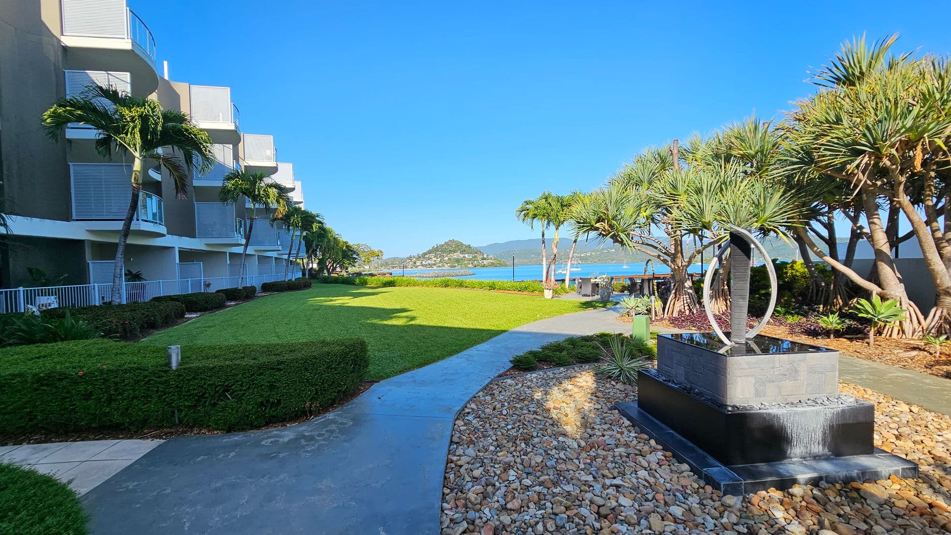 On the left, there is a modern building with balconies and palm trees lining the walkway. The middle features a well-maintained lawn leading to a view of a distant hill and water. On the right, a decorative water feature with a circular sculpture is surrounded by pebbles and more palm trees. The sky is clear and blue.