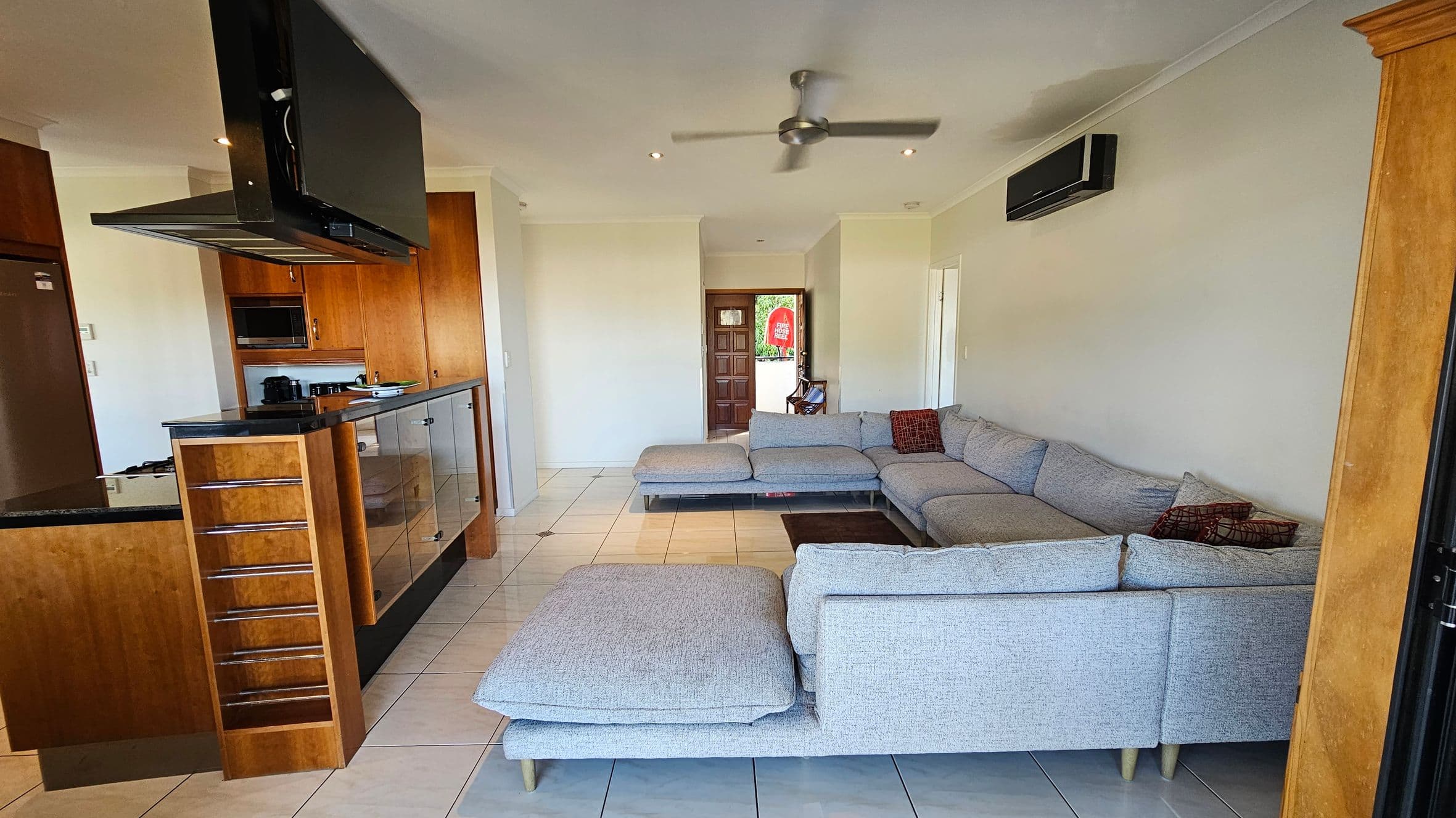 A modern living space with a large grey L-shaped sofa on the right, adorned with patterned cushions. The left side features a kitchen area with wooden cabinets, a built-in oven, and a black countertop. A ceiling fan is centrally located above the tiled floor. In the background, a wooden door with glass panels leads outside, and an air conditioning unit is mounted on the wall above the sofa.