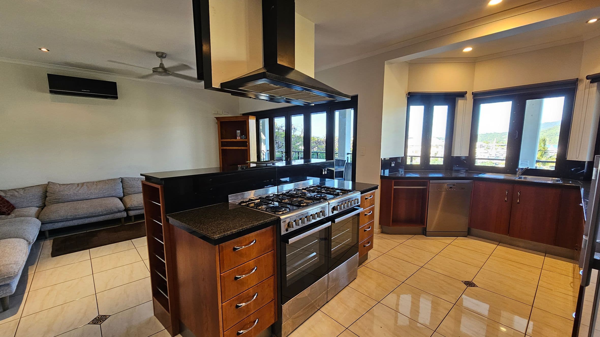 Modern kitchen and living area with a large stainless steel cooker and extractor hood in the centre. The kitchen features dark countertops and wooden cabinets. On the right, there are multiple windows with views of greenery. A dishwasher is integrated into the cabinetry. On the left, a grey sectional sofa sits against the wall beneath an air conditioning unit. The floor is tiled in a light colour, and the ceiling has recessed lighting and a ceiling fan.