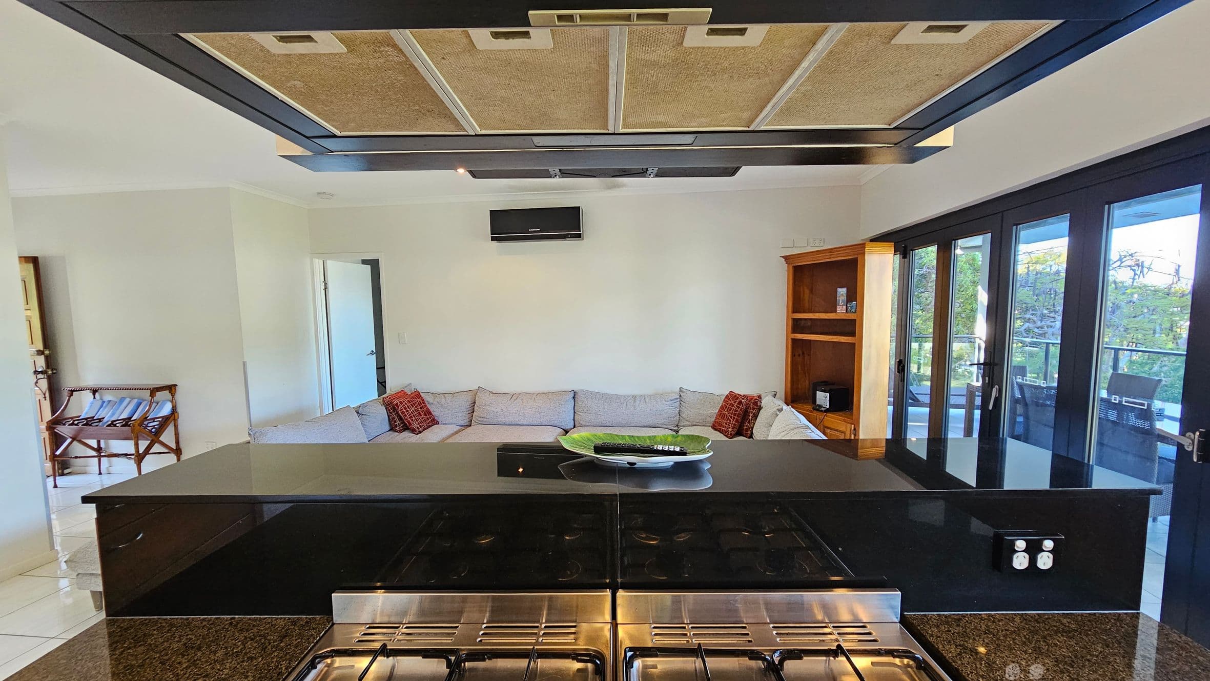 A modern kitchen with a black granite countertop in the foreground, featuring a gas stove. Above, a large extractor fan is visible. In the middle, a spacious living area with a light grey L-shaped sofa adorned with red cushions. A wooden shelving unit is on the right, next to large glass doors leading to a balcony with outdoor furniture. On the left, a small wooden luggage rack is near an open doorway. The walls are light-coloured, and the floor is tiled.