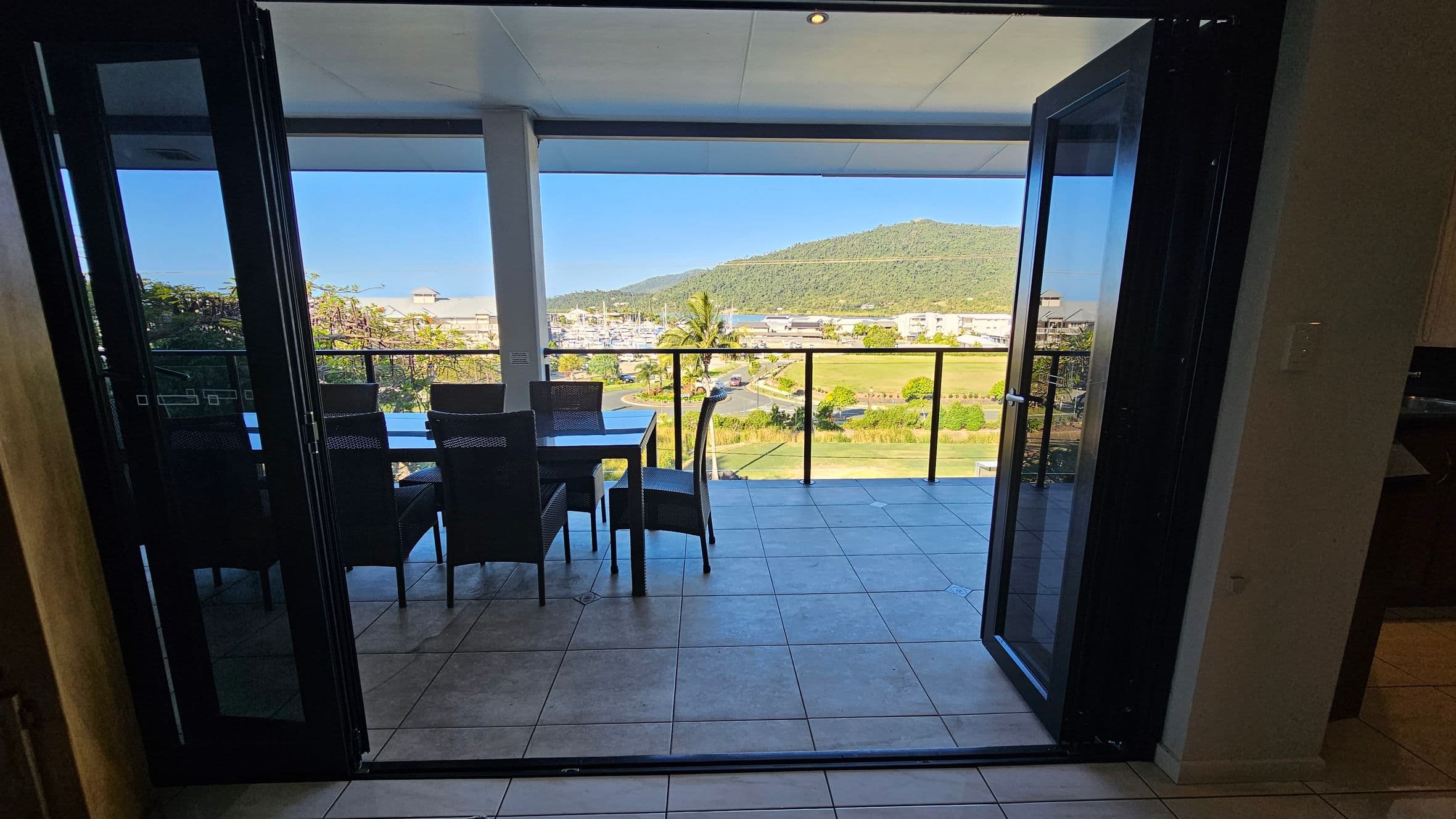 A view from inside a room looking out through open glass doors onto a tiled balcony. On the balcony, there is a dining table with several chairs. Beyond the balcony, a scenic view includes a lush green hill, a marina with boats, and a few buildings. The ceiling is smooth and white, and the floor inside is tiled. The balcony is enclosed with a glass railing.