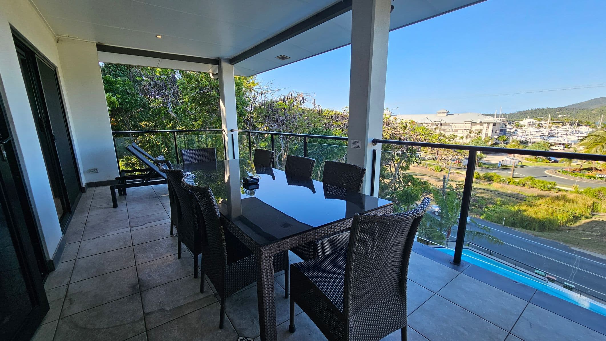 A balcony with a tiled floor and a glass-top dining table surrounded by wicker chairs. On the left, there are sliding glass doors leading inside. The balcony is bordered by a glass railing, offering a view of lush green trees and a marina with boats in the distance. The ceiling is white with recessed lighting.