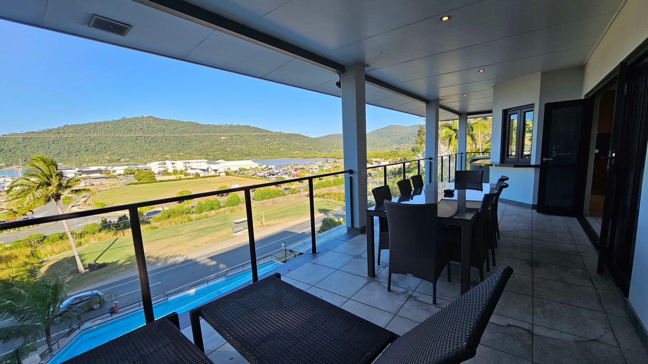 A spacious balcony with a tiled floor and a ceiling with recessed lights. On the right, there's a dining table with several wicker chairs. The left side features a glass railing offering a view of a road, a grassy area, and a marina with boats. In the background, lush green hills are visible. A palm tree is near the road, and a swimming pool is partially visible below. The balcony has large windows and a door leading inside.