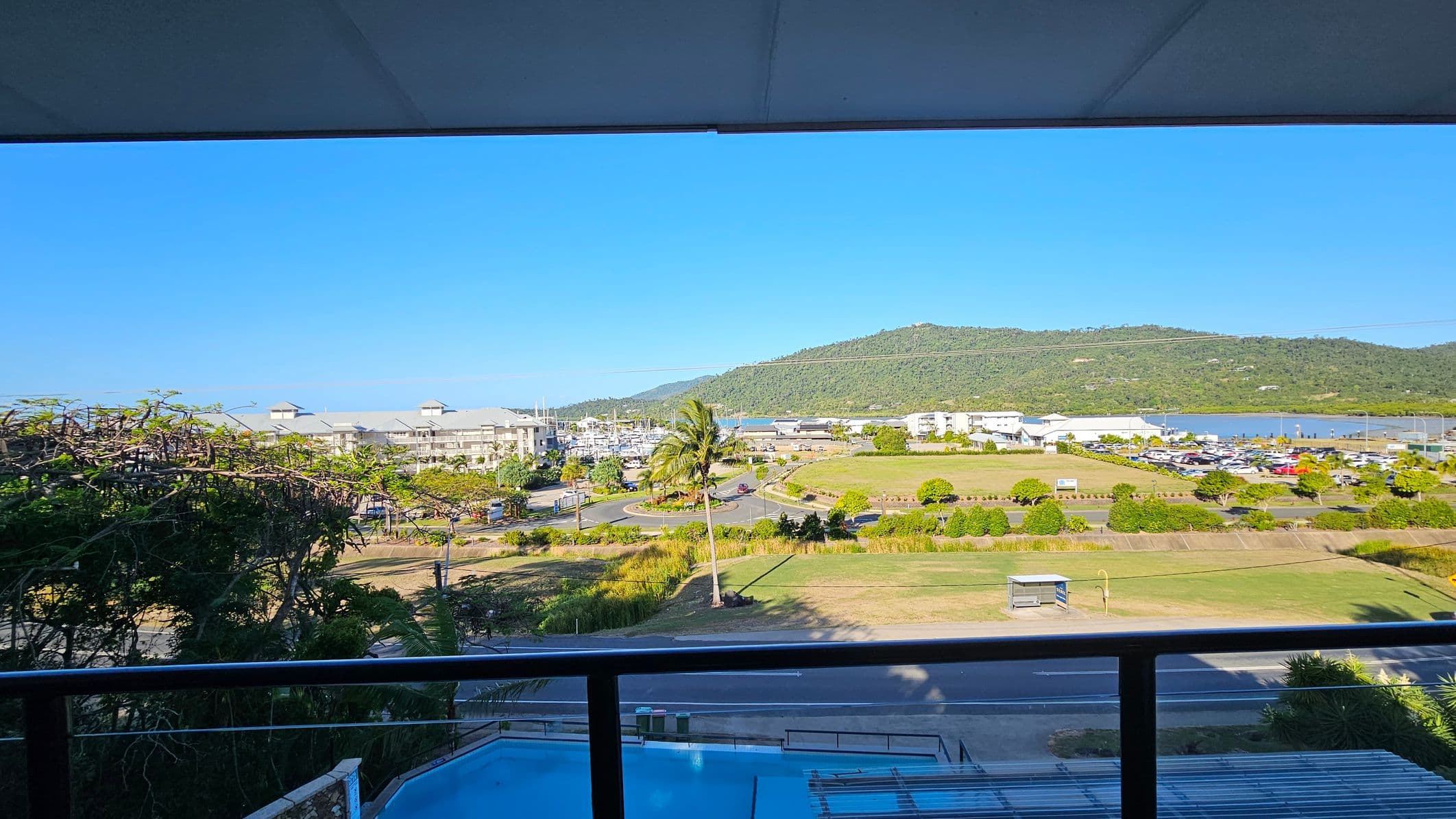 A view from a balcony with a glass railing in the foreground. Below, a road and a grassy area with a lone palm tree. In the middle distance, a marina with numerous boats and a cluster of buildings. Beyond, a green hill stretches across the background. On the left, dense foliage partially obscures the view. The ceiling of the balcony is visible at the top of the image.