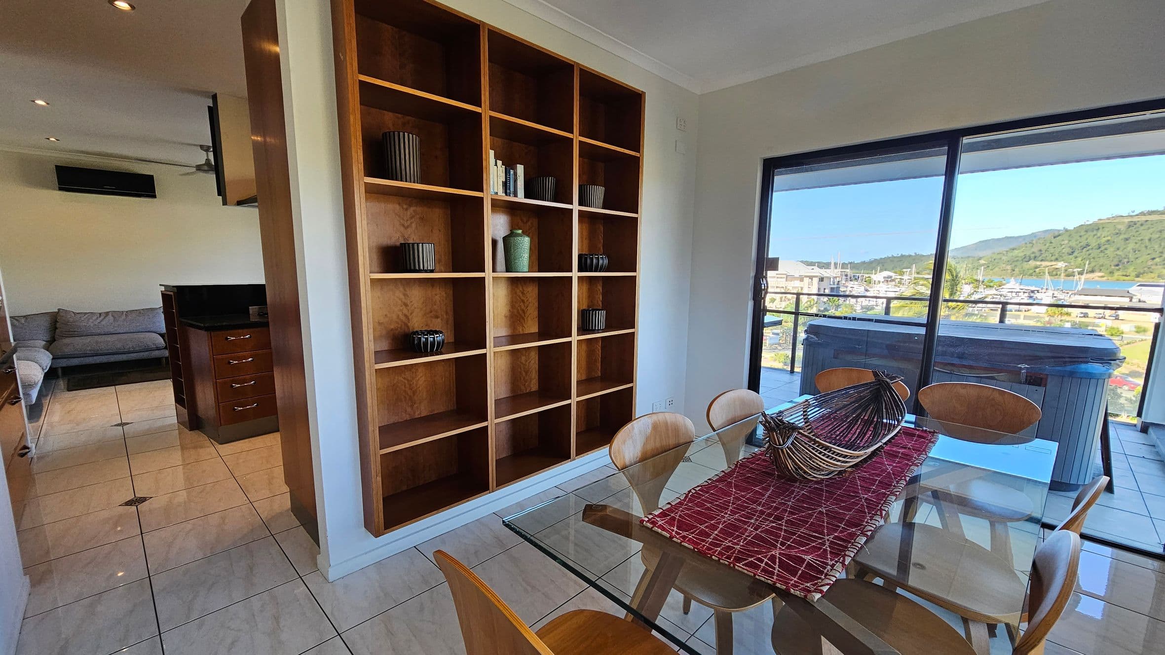 A room with a glass dining table and wooden chairs on the right, adorned with a red table runner and a decorative metal basket. Behind the table, large sliding glass doors open to a balcony with views of a marina and hills. On the left, a wooden shelving unit holds various decorative items. In the background, a grey sofa and a kitchen area with dark countertops are visible. The floor is tiled in a light colour.
