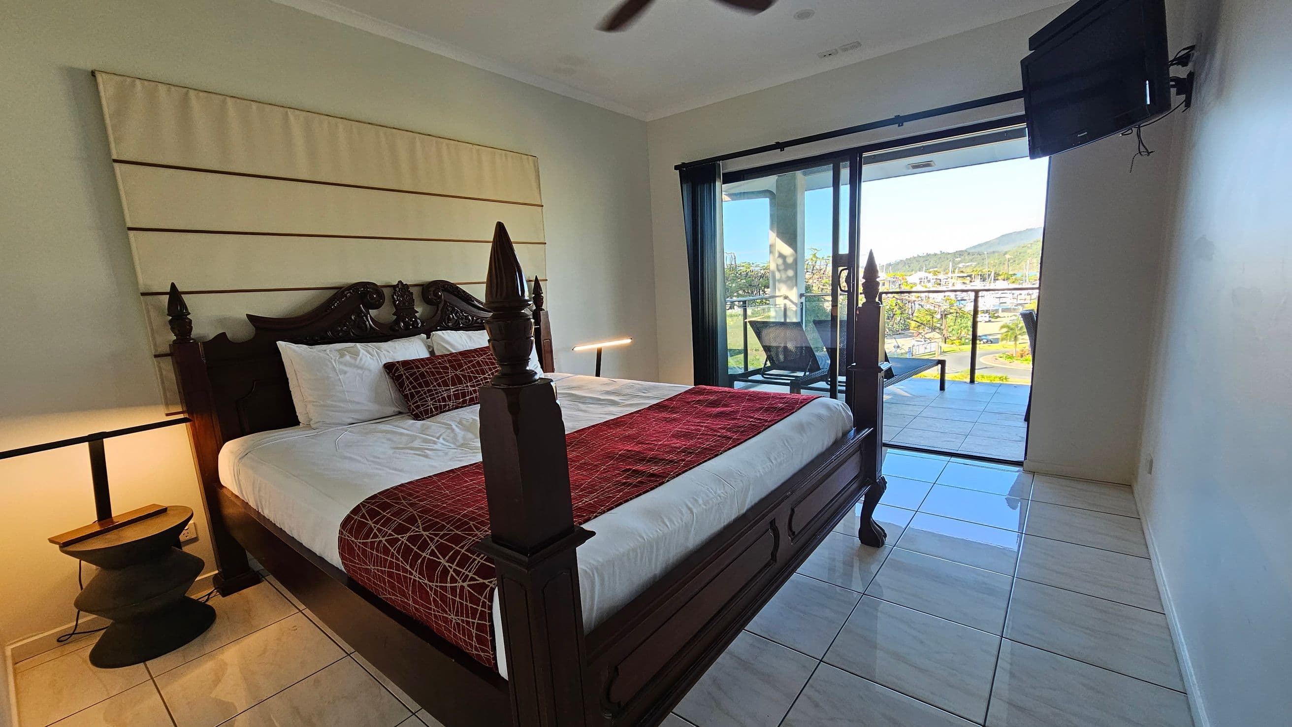 A bedroom with a large, dark wooden bed featuring an ornate headboard and posts. The bed is dressed with white sheets, a red patterned throw, and matching pillows. To the left, a modern black side table with a lamp. On the right, a wall-mounted TV. The room opens to a balcony with glass doors, showing outdoor chairs and a view of greenery and distant hills. The floor is tiled in a light colour, and the ceiling is plain white.