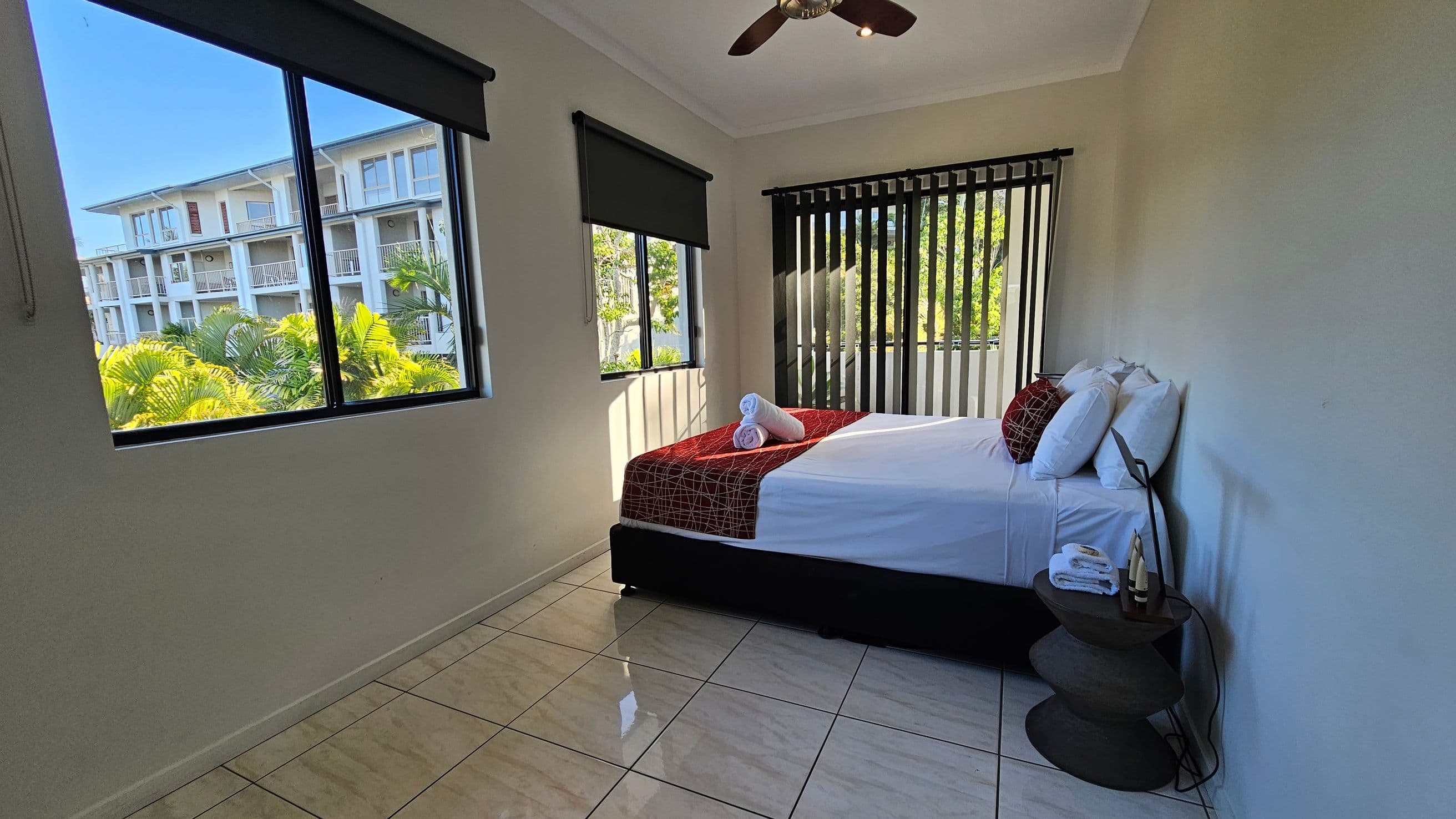 A bedroom with a bed on the right, covered with a red and white patterned throw and white pillows. Two rolled towels are placed on the bed. A small round bedside table holds folded towels and toiletries. The room has large windows with black blinds on the left and at the back, offering a view of a building and lush green trees. The floor is tiled in a light colour, and a ceiling fan is visible above.
