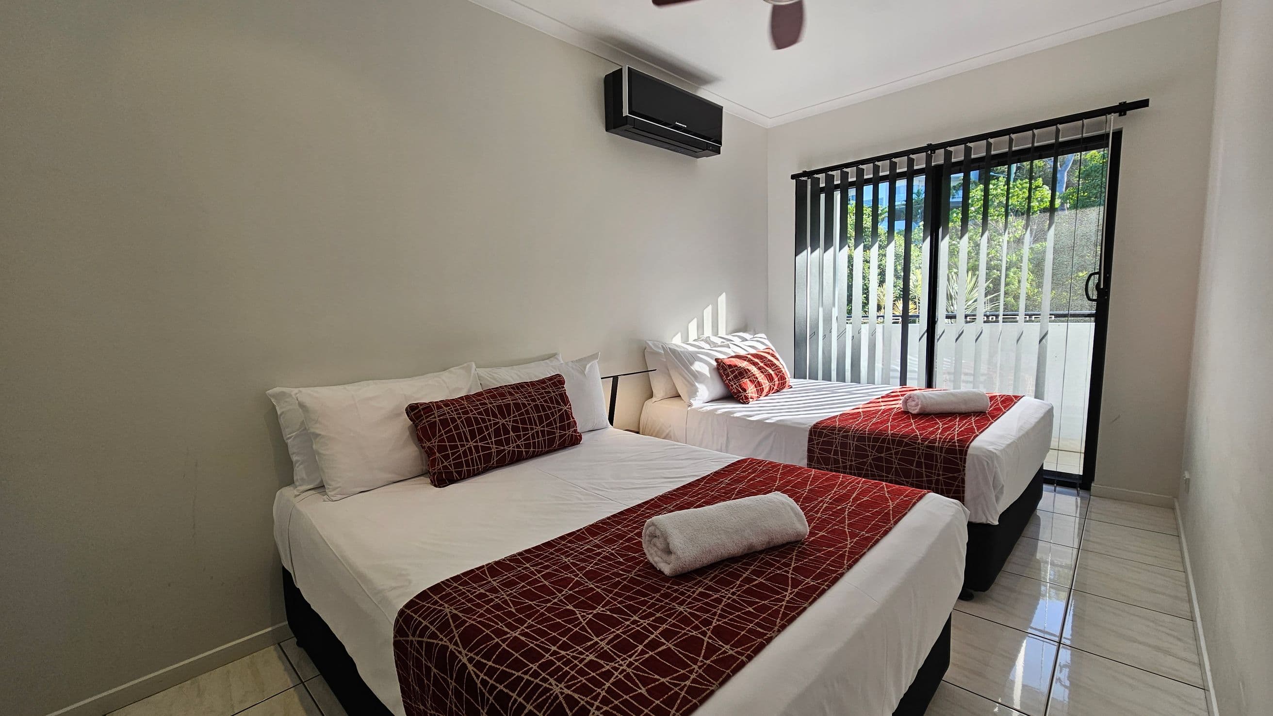 Two single beds with white sheets and red patterned throws are positioned side by side. Each bed has a rolled white towel and a matching red cushion. Above the beds, a black air conditioning unit is mounted on the wall. To the right, vertical blinds cover a sliding glass door, allowing sunlight to filter in. The floor is tiled in a light colour, and the ceiling is plain white. Outside, greenery is visible through the glass.