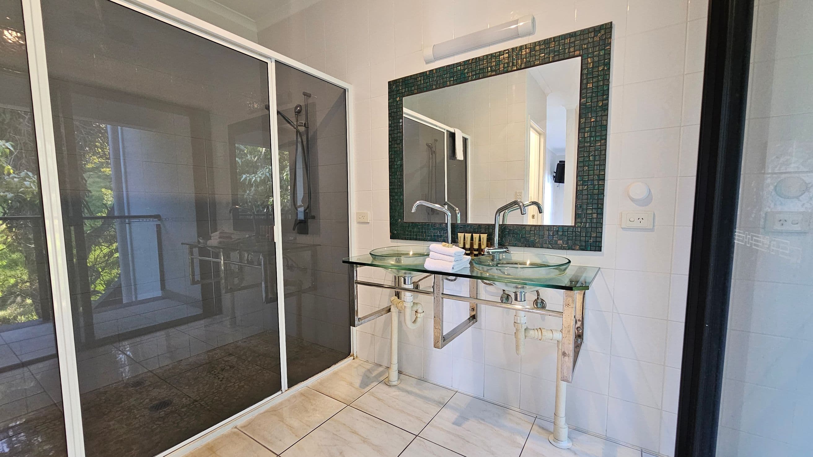 A bathroom with a large glass shower enclosure on the left, featuring a sliding door. On the right, there is a double sink vanity with glass basins and chrome fixtures. Above the sinks is a large mirror with a mosaic tile border. The floor is tiled in a light colour, and there are small toiletries and folded towels on the vanity. The walls are tiled in white, and there is a light fixture above the mirror.