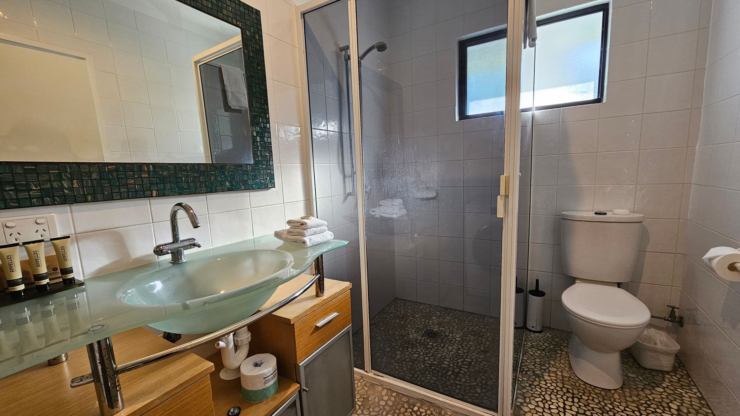 A bathroom with a glass sink and chrome tap on the left, set on a wooden cabinet with toiletries and a power outlet nearby. Above the sink is a large mirror with a dark mosaic frame. In the centre, a glass shower enclosure with a pebble floor and a small window. On the right, a white toilet with a roll of toilet paper on a holder. The floor is covered with pebble-like tiles, and the walls are tiled in white.