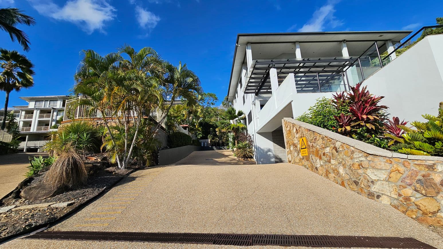 A paved driveway leads up to two modern buildings surrounded by lush greenery. On the left, a sign reads "Boathaven Bay Holiday Apartments" near a cluster of palm trees. On the right, a building with a sign "Waterfront Whitsunday Retreat" is partially visible, with a stone wall and landscaped garden in front. The sky is clear and blue.