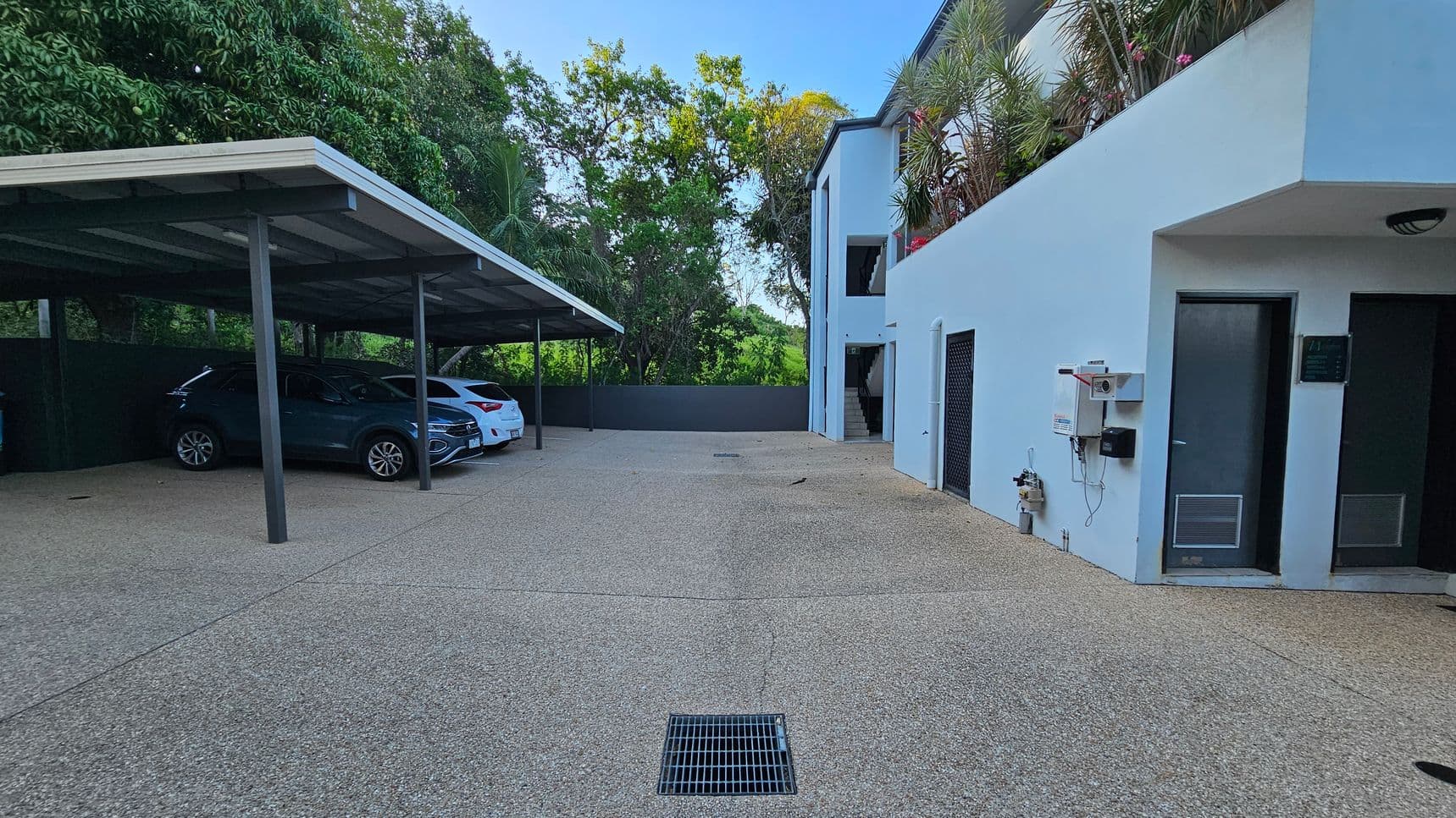 A car park area with a covered section on the left, sheltering two cars, one dark and one white. The ground is paved with a drainage grate in the centre. On the right, a light blue building with a door and some utility boxes attached to the wall. In the background, there are tall trees and greenery.