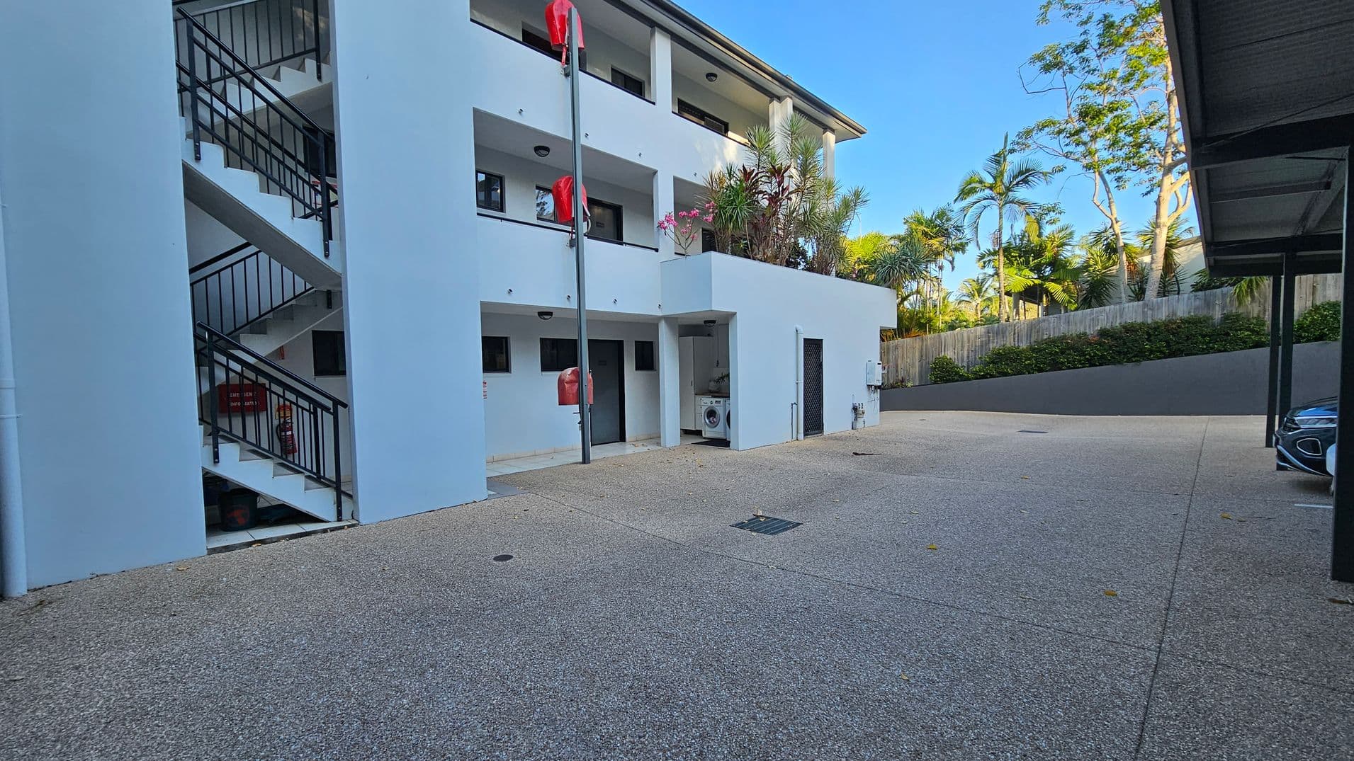 A three-storey building with white walls and black railings on the left, featuring an external staircase. Red fire hydrants are mounted on poles. The ground is paved with a textured surface. On the right, there's a covered parking area with a car partially visible. In the background, a wooden fence with lush green plants and trees adds a natural element. A washing machine is visible in an alcove on the ground floor.