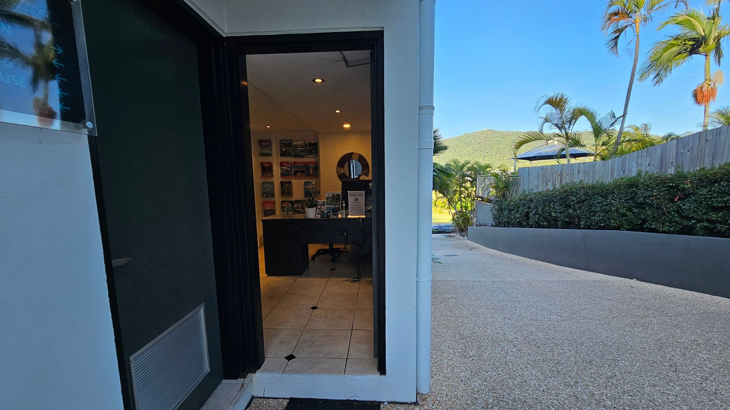 An open doorway on the left leads into a room with a desk, brochures on the wall, and a circular mirror. The floor inside is tiled with a light pattern. Outside, on the right, there's a gravel path bordered by a low hedge and a wooden fence. Palm trees and a distant hill are visible in the background. The exterior wall is light-coloured, and the ceiling inside has recessed lighting.