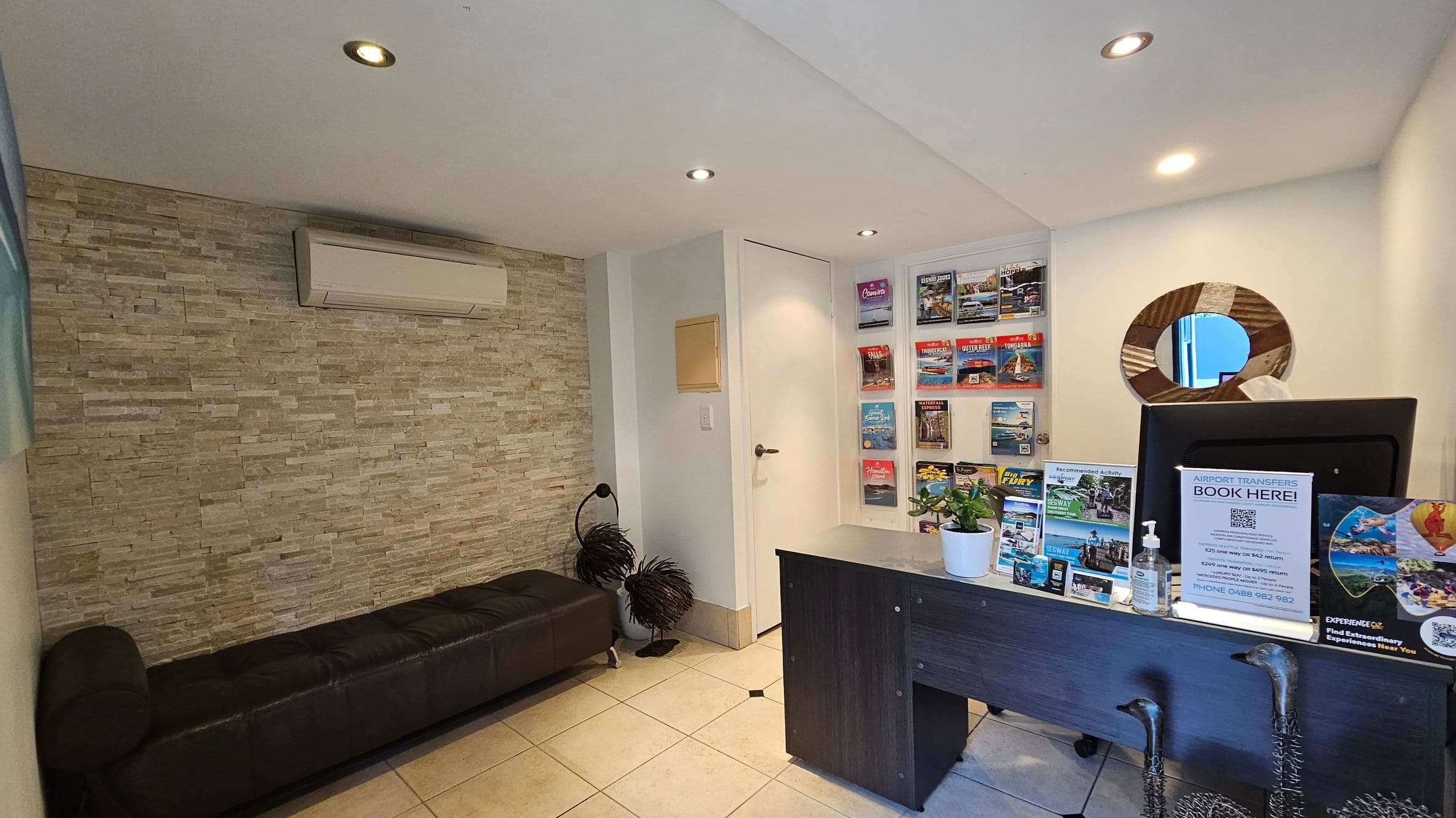 A small office space with a stone-textured wall on the left, featuring a black leather bench and an air conditioning unit above. On the right, a dark wooden desk displays brochures, a potted plant, and a hand sanitiser bottle. Behind the desk, a wall-mounted display holds various colourful brochures. A round mirror with a wooden frame is on the wall above the desk. The floor is tiled, and the ceiling has recessed lighting.