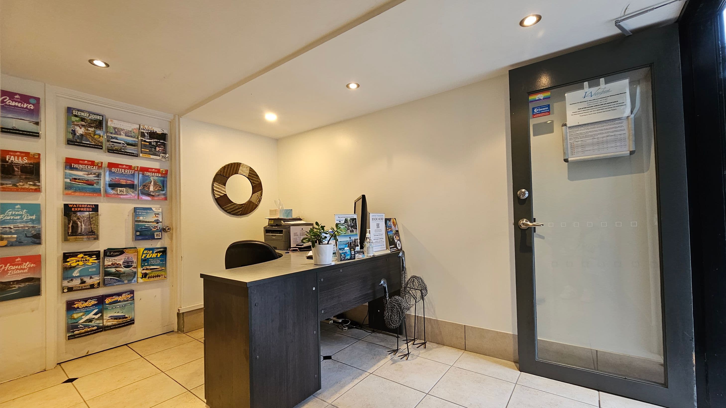 A small office space with a dark wooden desk on the right, featuring a computer monitor, potted plant, and brochures. Behind the desk is a circular decorative wall piece. On the left wall, there are multiple travel brochures displayed in a grid. The floor is tiled in light beige, and the ceiling has recessed lighting. A glass door with notices is on the far right.