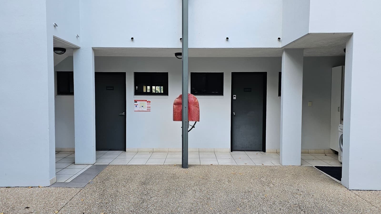 A light blue building facade with two dark doors and small rectangular windows above them. A red fire hose reel is mounted on a central grey pole. The floor is a mix of gravel and tiles, with some fallen leaves scattered. The ceiling is flat and white, with a light fixture visible. On the right, a glimpse of a washing machine is seen through an open area.