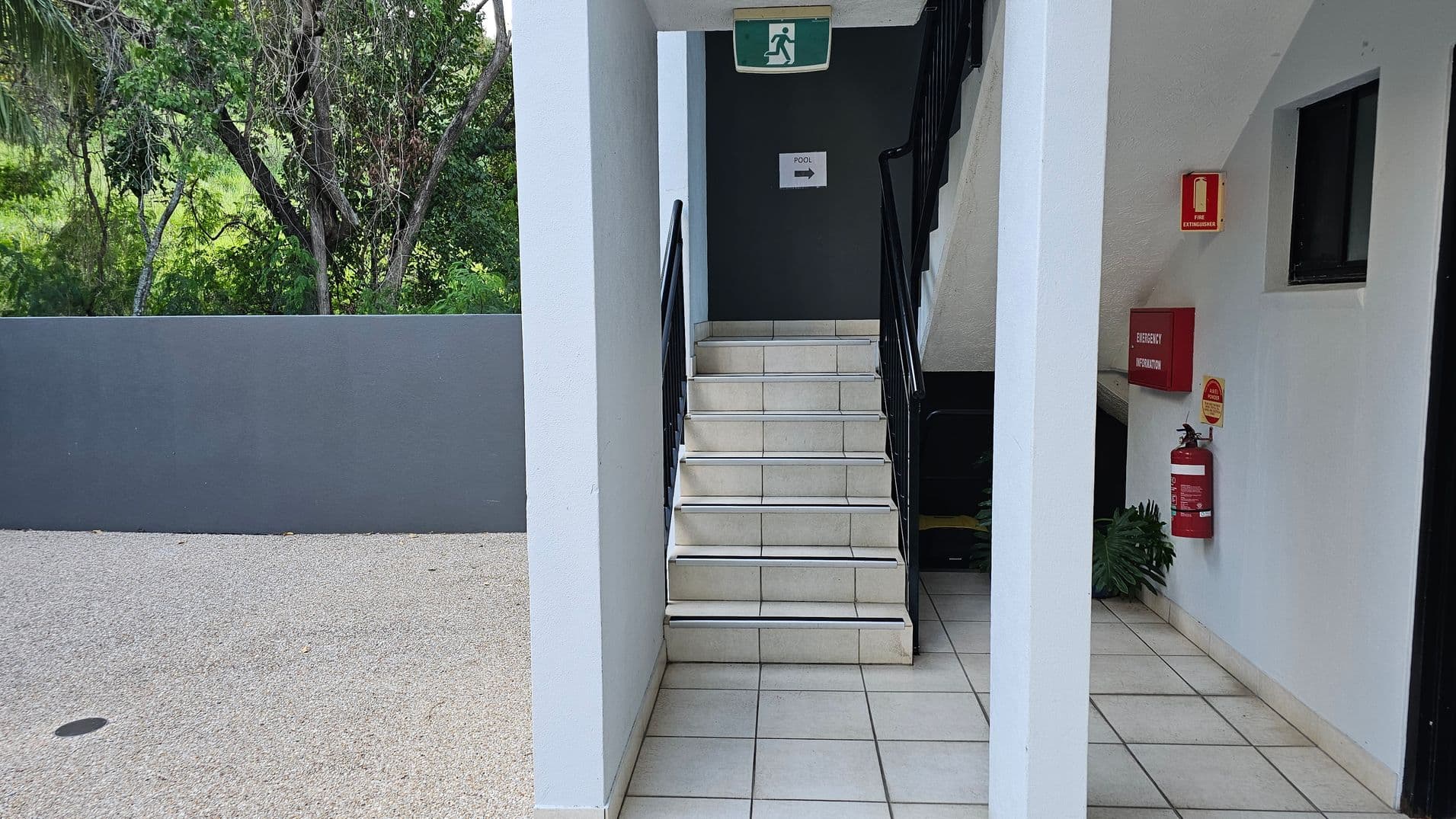A stairway with white tiles and black railings is in the centre, leading upwards. Above the stairs is an emergency exit sign. To the right, a fire extinguisher and an emergency information box are mounted on the wall. A small window is also visible. On the left, a grey wall separates the area from a green, tree-filled landscape. The floor is tiled, and the ceiling is white.