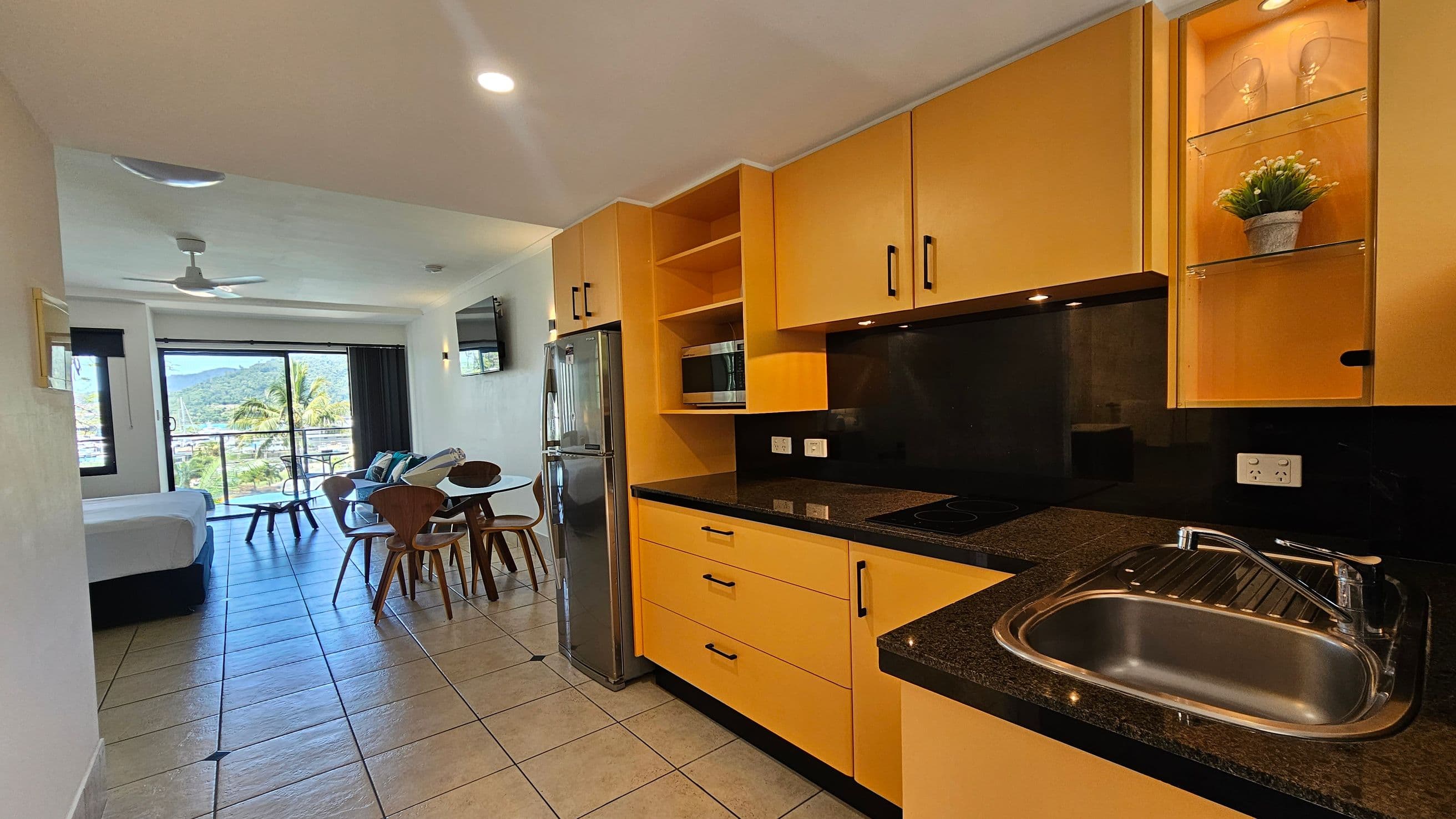 A modern kitchen and living area with light-coloured tiled flooring. On the right, there are yellow cabinets with black countertops, a sink, and a built-in microwave. A stainless steel fridge is next to the cabinets. On the left, a dining table with wooden chairs is visible, leading to a bed near a large window. The window offers a view of palm trees and distant hills. A ceiling fan and a wall-mounted TV are also present.