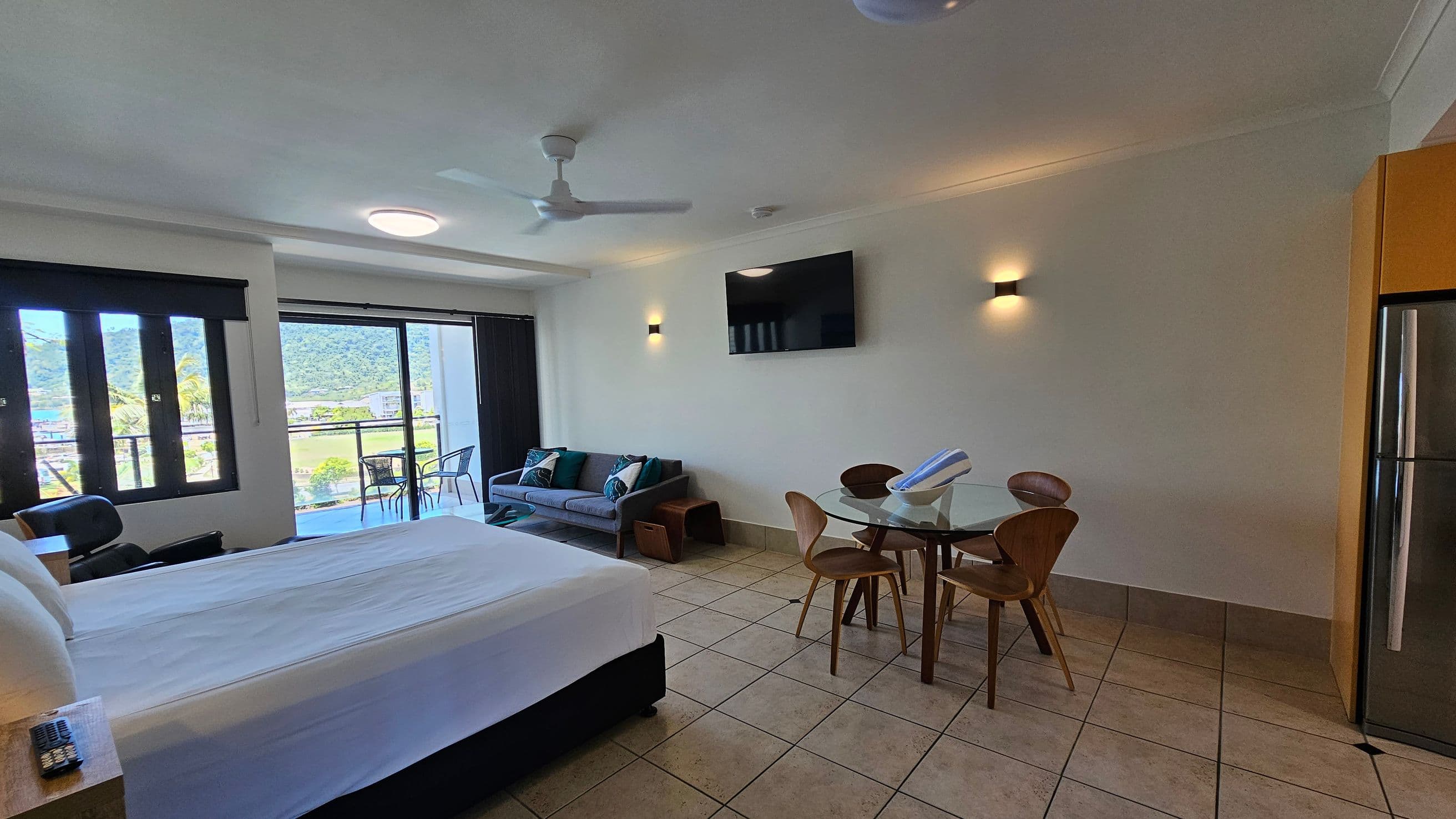 A modern hotel room with a large bed on the left, facing a wall-mounted TV. In the centre, there's a glass dining table with four wooden chairs. A grey sofa with decorative cushions is positioned against the wall, near a sliding glass door leading to a balcony with outdoor chairs. The floor is tiled, and the ceiling features a fan and recessed lighting. A stainless steel fridge is on the right.